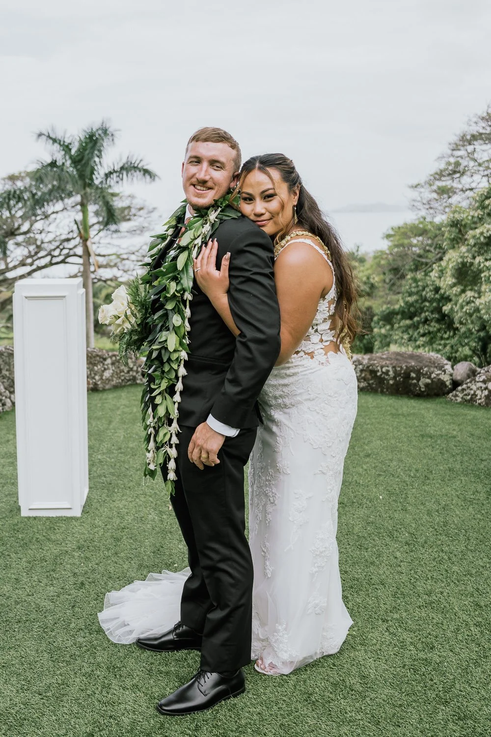 A newlywed couple embracing outdoors on a grassy area with trees and a body of water in the background. The bride is wearing a white lace wedding dress and the groom is in a black suit with a floral lei.