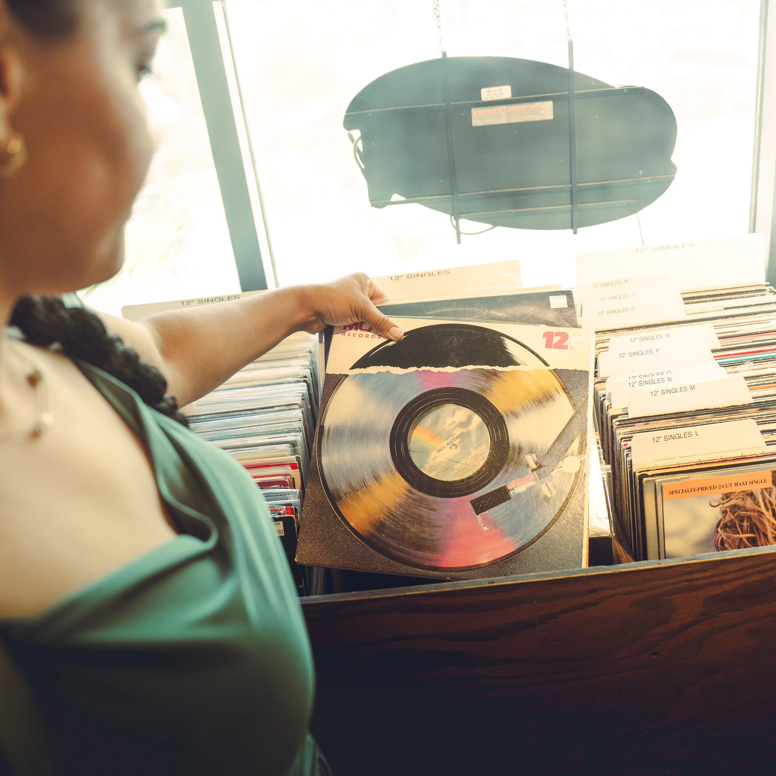 A person with braided hair wearing a necklace is looking at a display of vinyl records in a store. The records are organized in rows with visible labels indicating their sizes and types, such as "12" SINGLES." The person's hand is reaching toward the