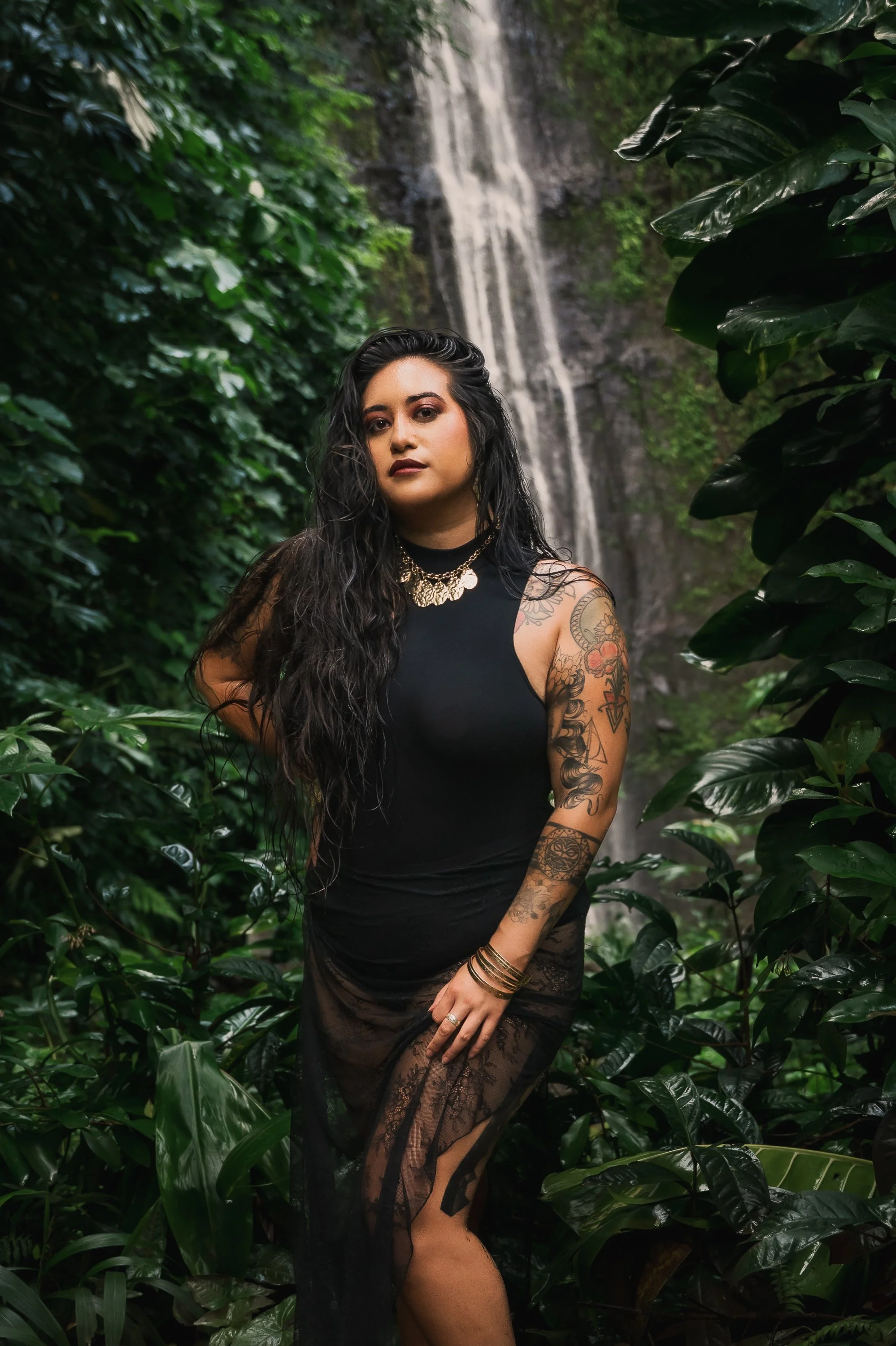 A woman with long dark hair, wearing a black sleeveless dress with lace details, standing among lush green plants with a waterfall in the background.