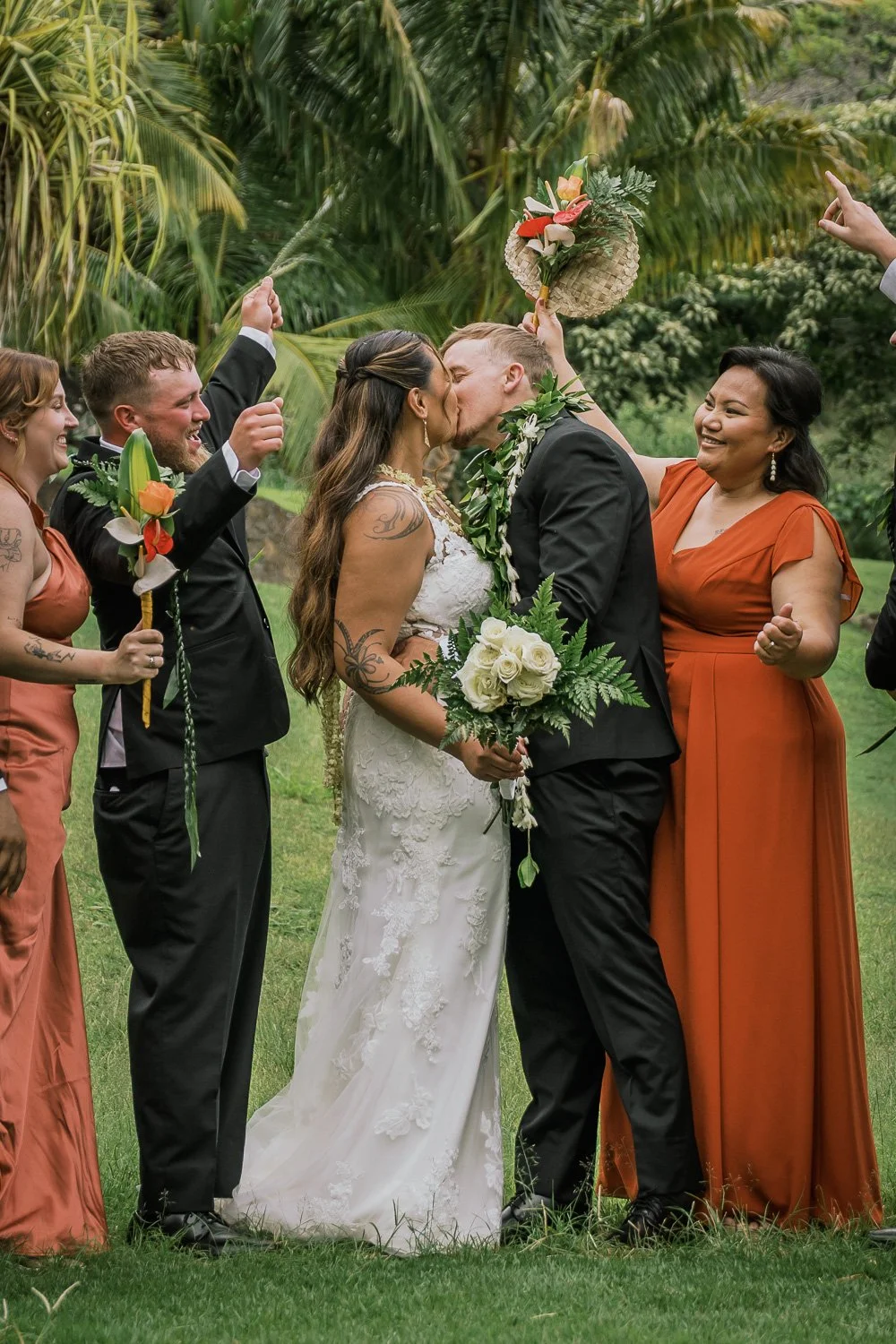 A bride and groom kiss during their wedding ceremony outdoors, surrounded by family and friends, with lush green trees in the background.