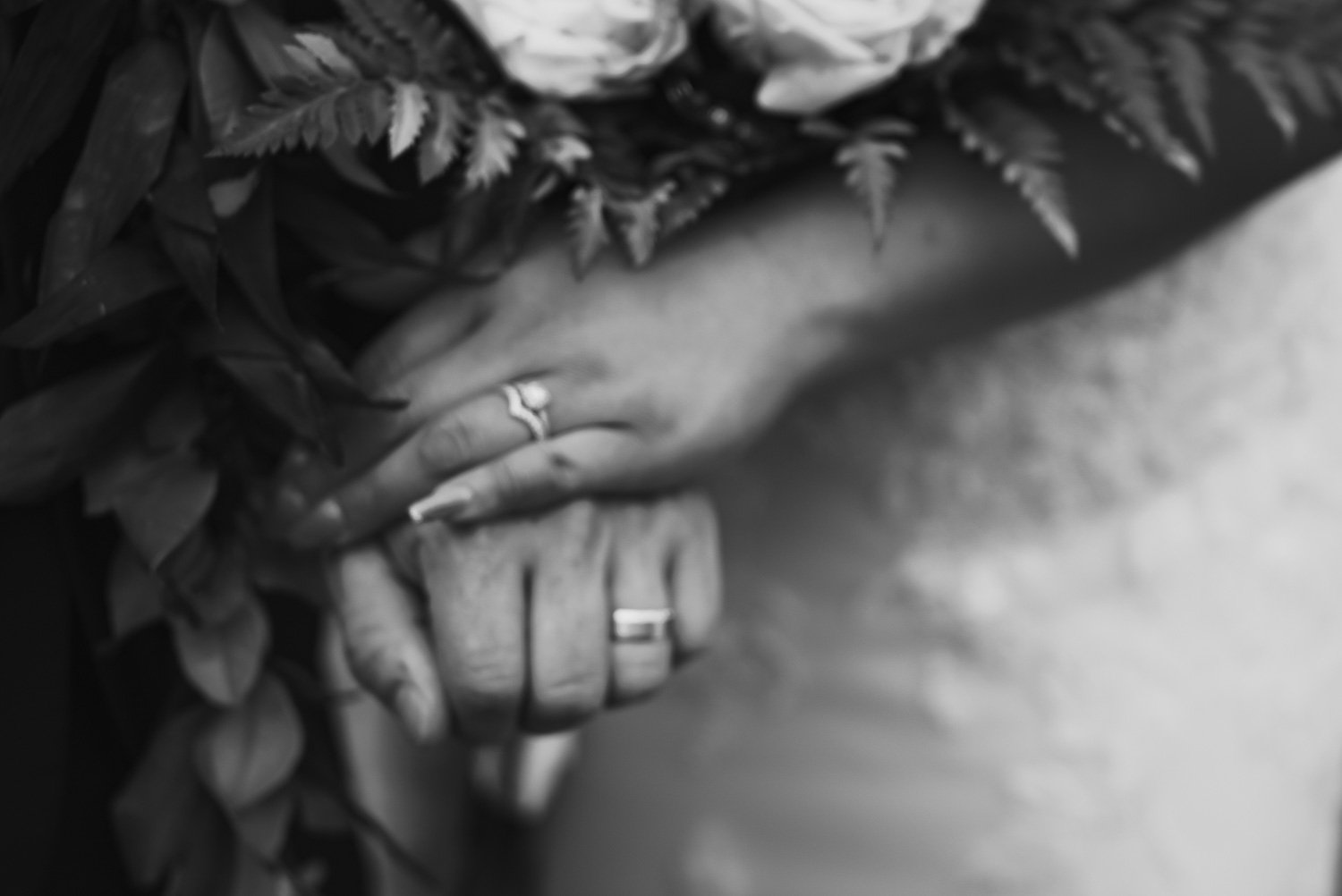 Close-up of two hands gently clasped, with rings on the fingers, surrounded by foliage and flowers in a black-and-white photograph.
