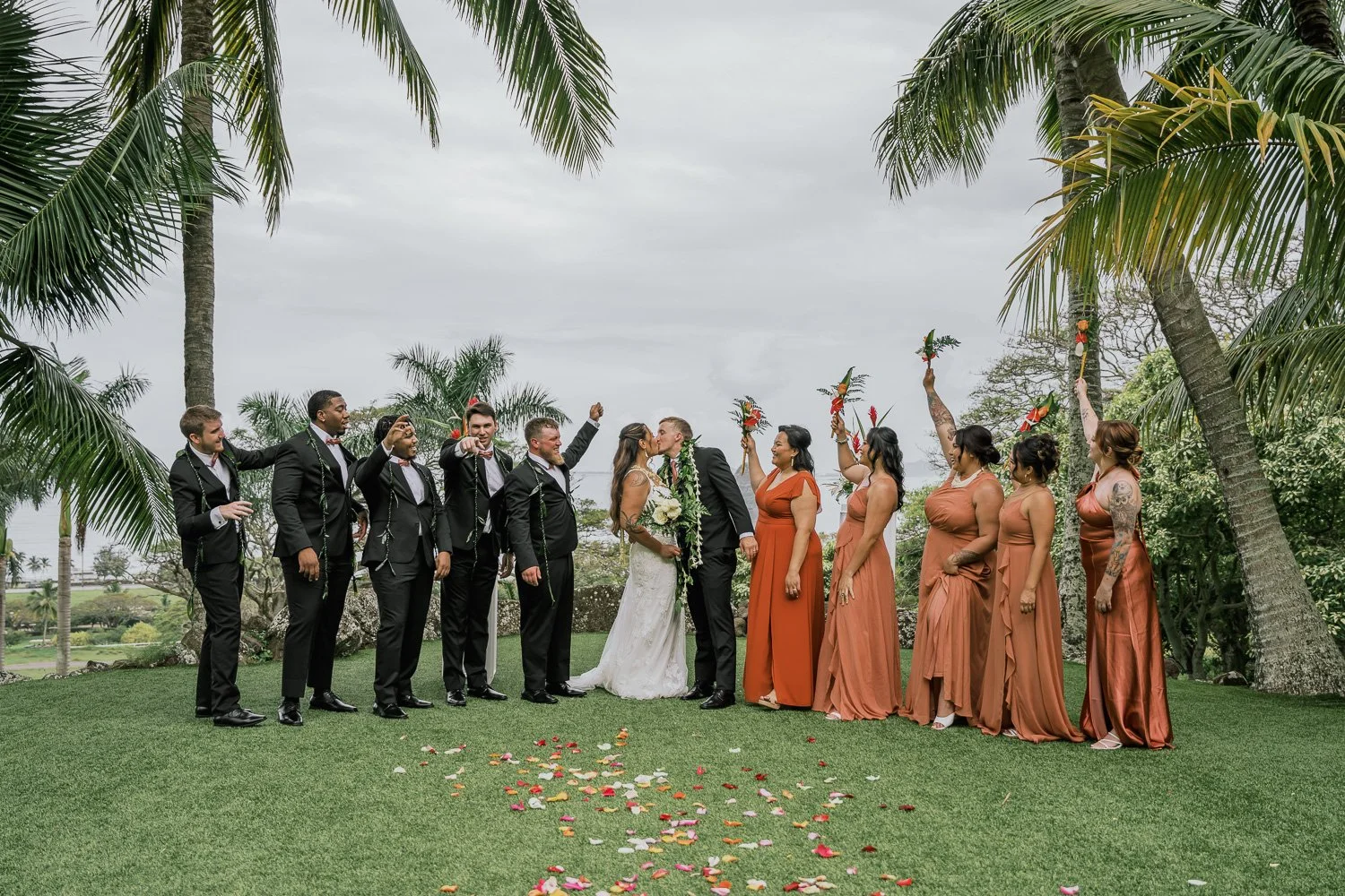 A wedding ceremony outdoors with a bride and groom kissing, surrounded by wedding party members and bridesmaids holding floral arrangements, on a grassy area with palm trees, overcast sky, colorful flower petals on the ground, and lush greenery in th