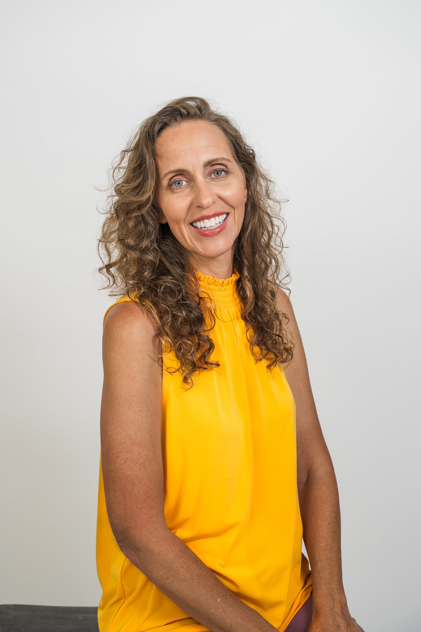 A woman with curly brown hair wearing a sleeveless yellow top, smiling in front of a plain white background.