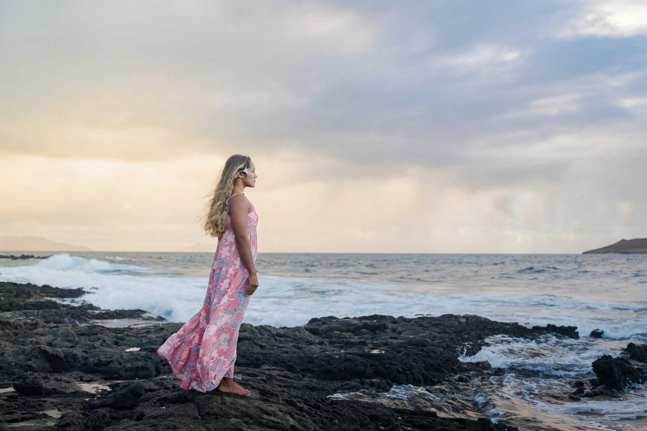 A woman in a pink floral dress stands barefoot on black rocks at the edge of the ocean during sunset, looking out at the sky and water.