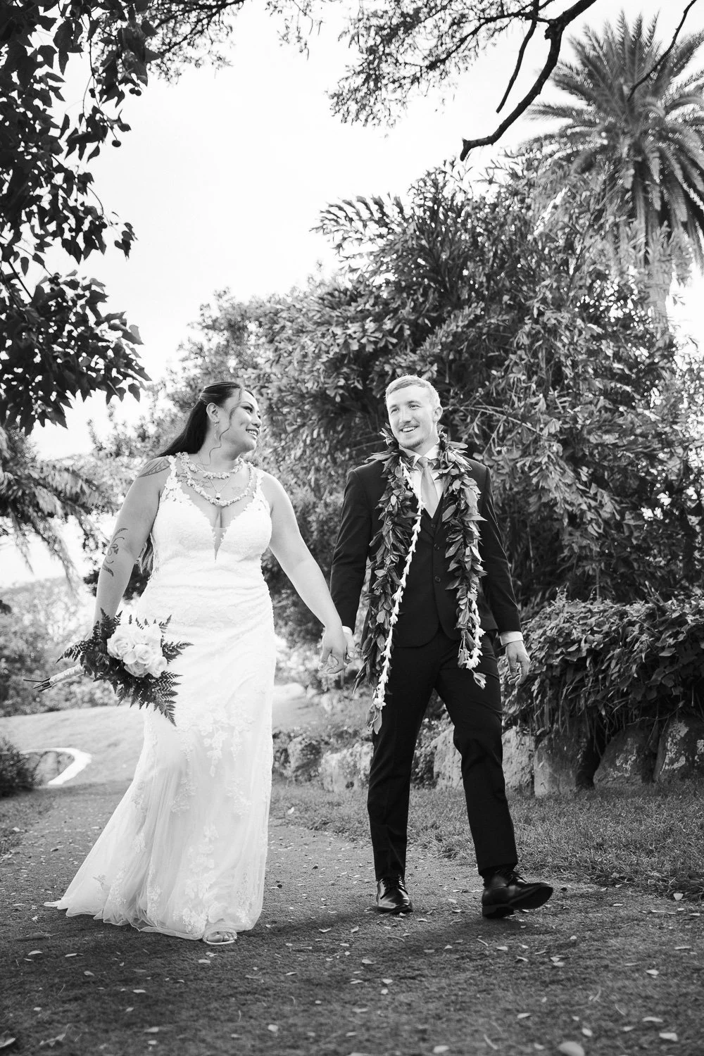 A black and white photo of a wedding couple walking outdoors, holding hands, surrounded by trees and lush foliage. The bride wears a lace wedding gown and holds a bouquet, while the groom is dressed in a suit with a lei around his neck, both smiling.