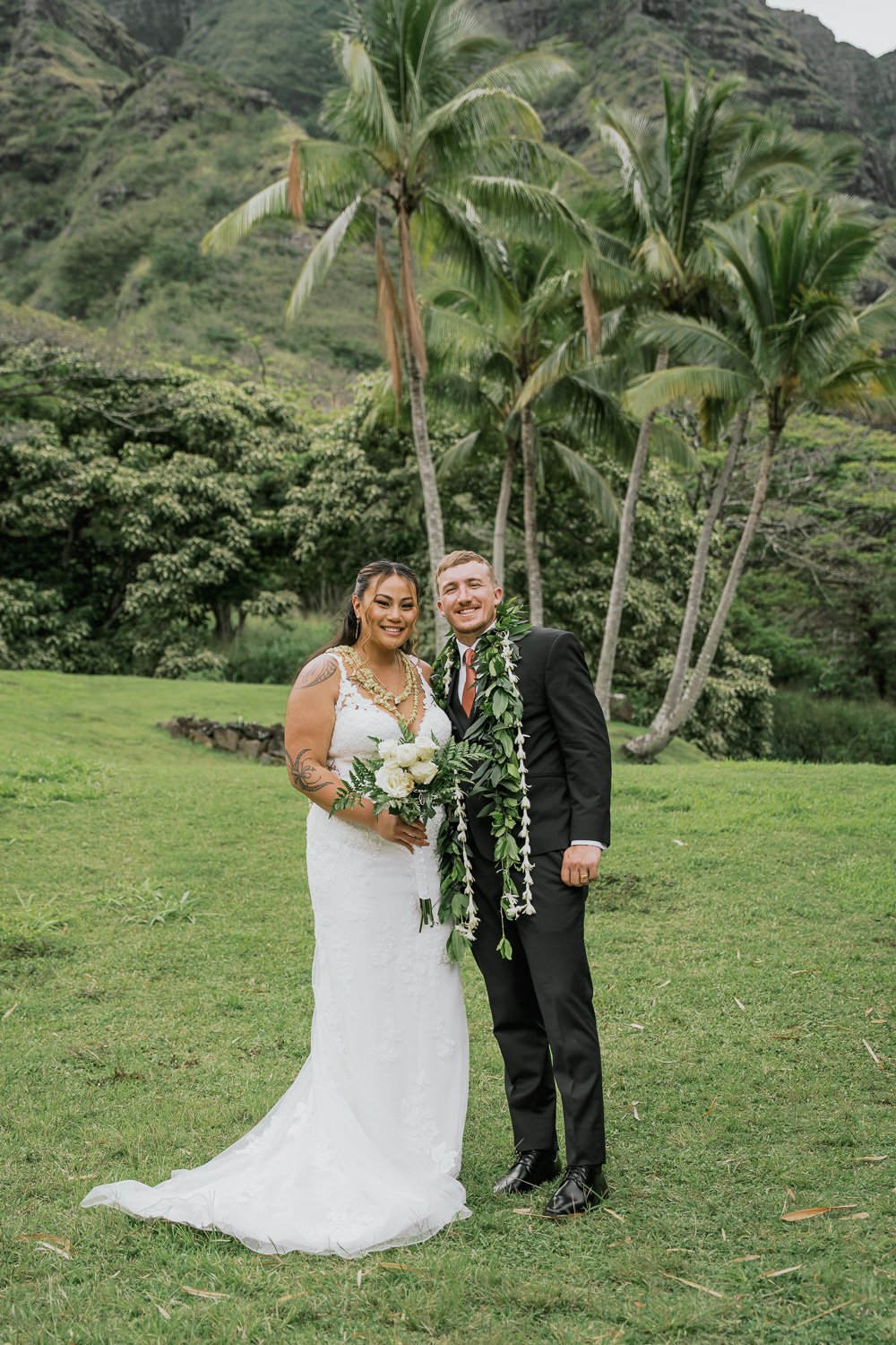 A couple dressed in wedding attire standing outdoors on a grassy area with tropical trees, including palm trees, and mountains in the background.