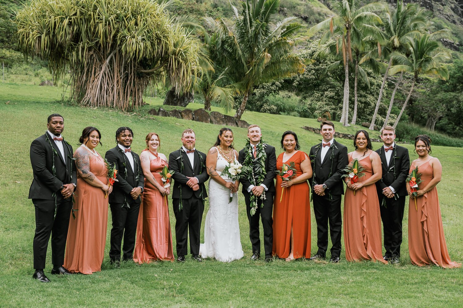 Group of people at outdoor wedding, including bride in white gown and groom in black suit with leis, surrounded by bridesmaids and groomsmen in formal attire, standing on green grass with tropical trees in the background.