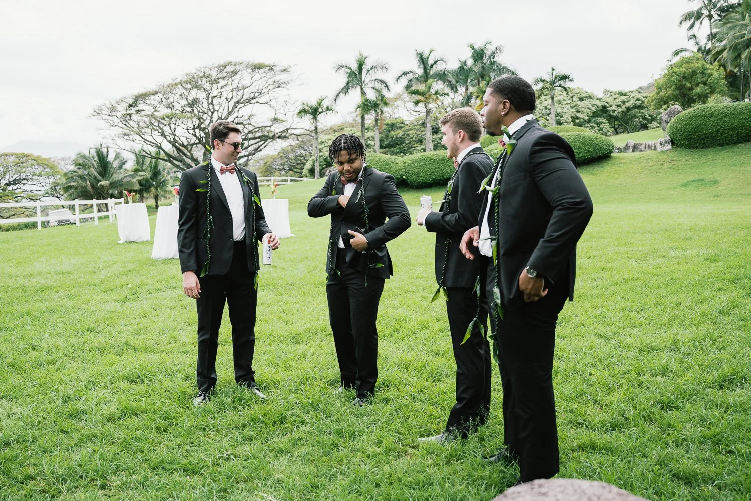 Group of five men dressed in black suits and ties standing on a grassy lawn, engaging in conversation at an outdoor event with green trees and shrubs in the background.