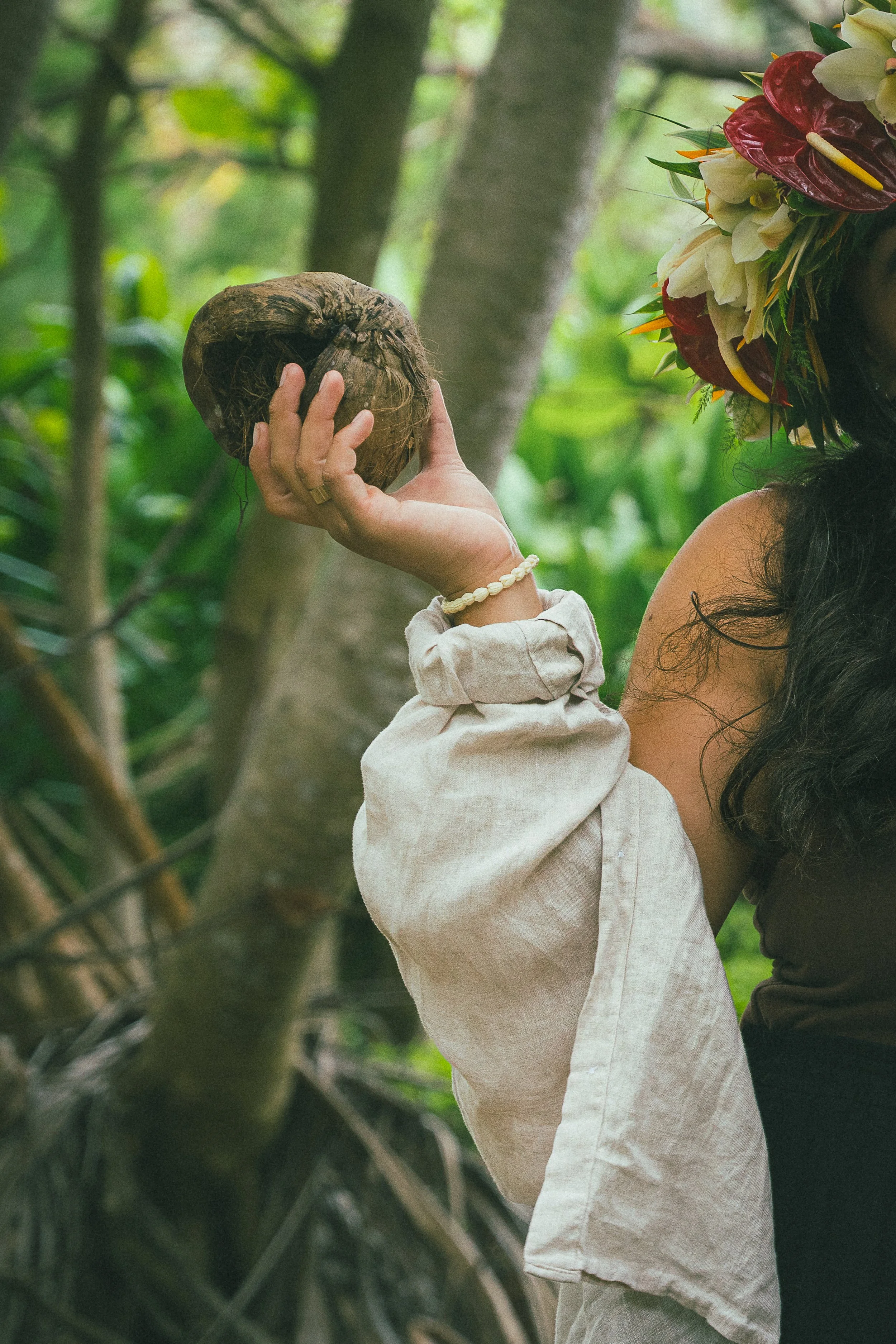 A woman holding a coconut in a lush green area, with a child wearing a beige shirt and a shell bracelet, wearing a floral crown, standing near her.