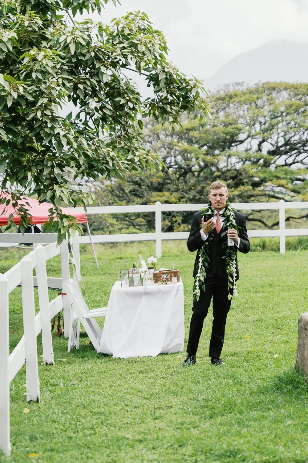 A man in a black suit with a pink tie and a green leafy lei around his neck stands outdoors on a grassy area, holding a drink in one hand and a smartphone in the other. There is a white fenced area with a small white table covered with a white cloth,