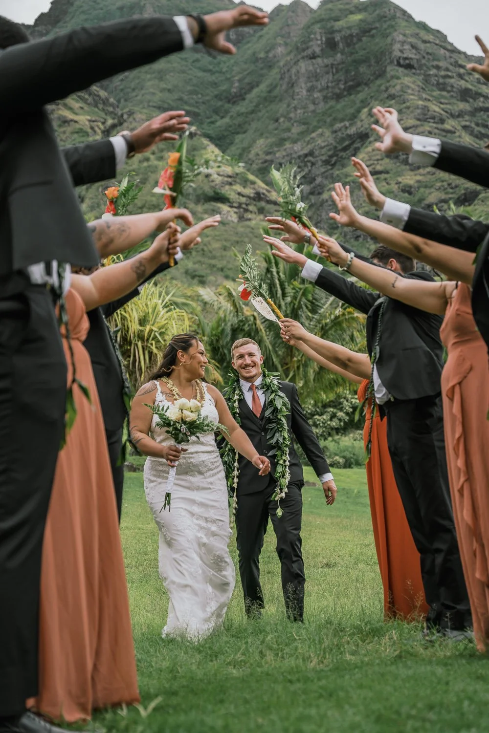 A bride and groom holding hands and smiling as they walk through a floral tunnel created by friends and family holding up decorated branches and greenery in an outdoor wedding setting with lush mountains in the background.