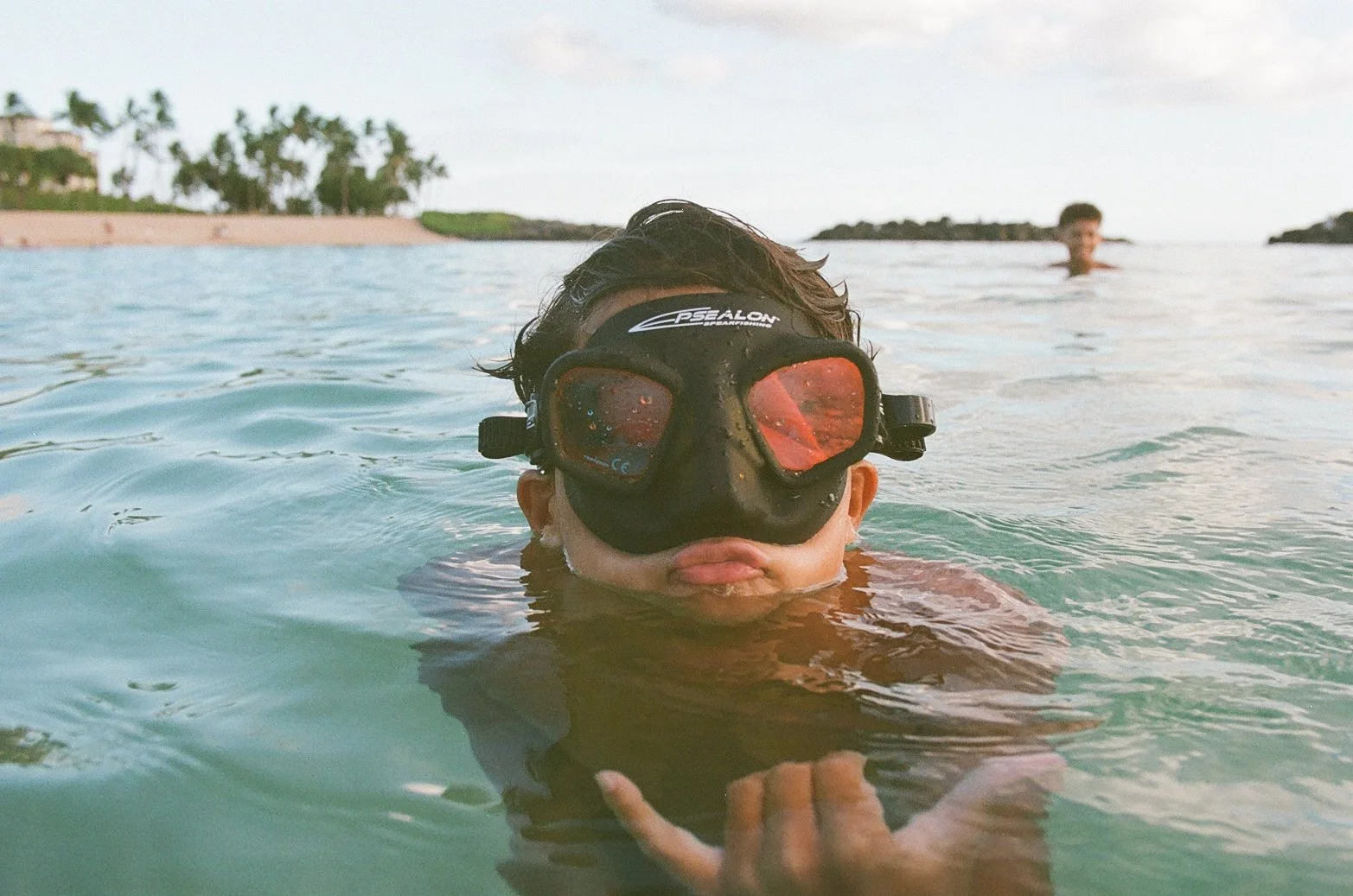 A person wearing a black diving mask with red lenses, partially submerged in water, with a background of a beach and another person swimming in the distance.