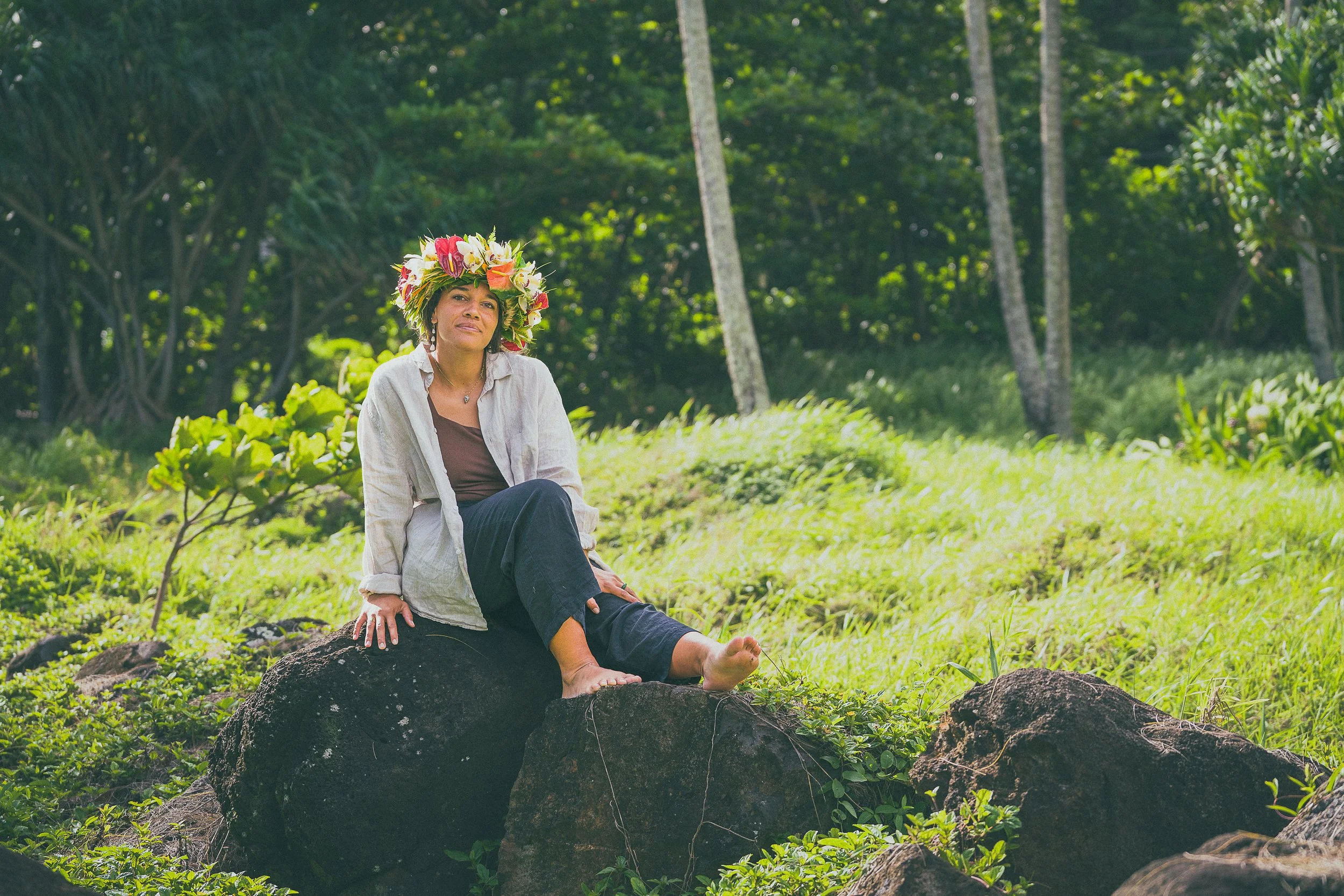 A woman sitting on a large rock in a lush, green forest, wearing a floral crown and casual clothing.