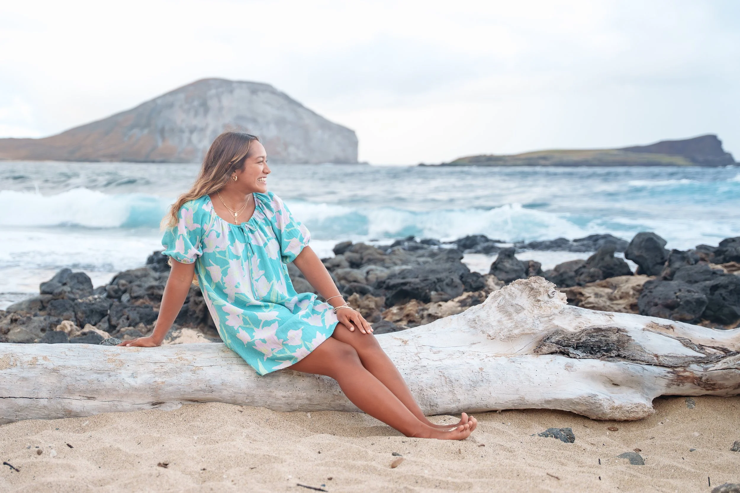 A woman sitting on a large piece of driftwood on a sandy beach, with dark rocks and the ocean in the background, smiling and looking to her right.