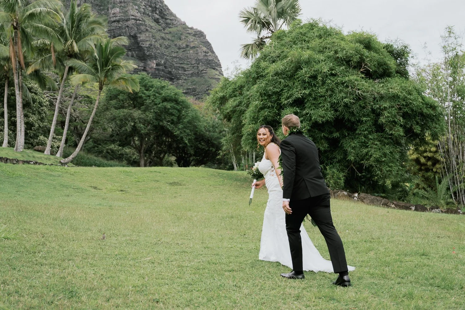 A wedding couple walking on a green lawn in a tropical garden, with tall palm trees and mountainous terrain in the background. The bride is smiling while holding a bouquet, and the groom is dressed in a black suit.