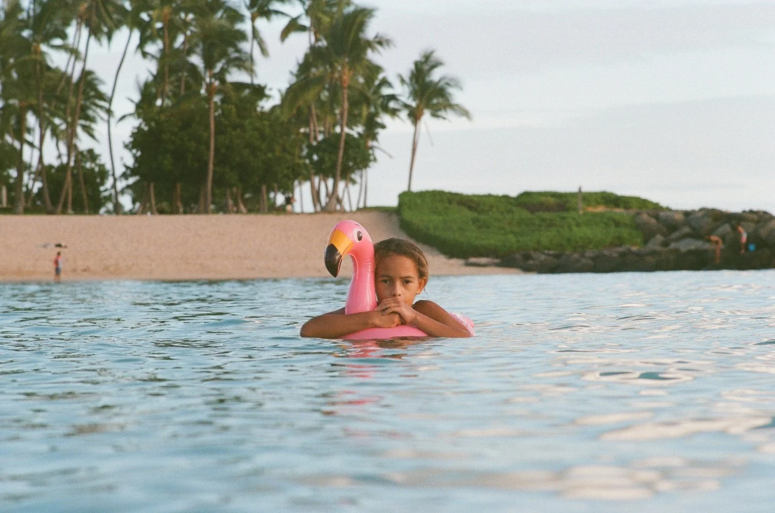 A young girl in a pink flamingo float rests in the water at the beach, with a background of palm trees, sandy shore, and rocky outcrop.