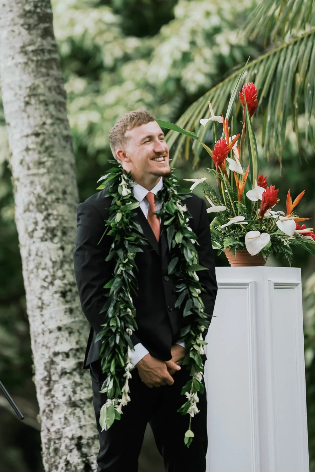 A groom in a black suit and lei stands under trees during a wedding in a lush outdoor setting.