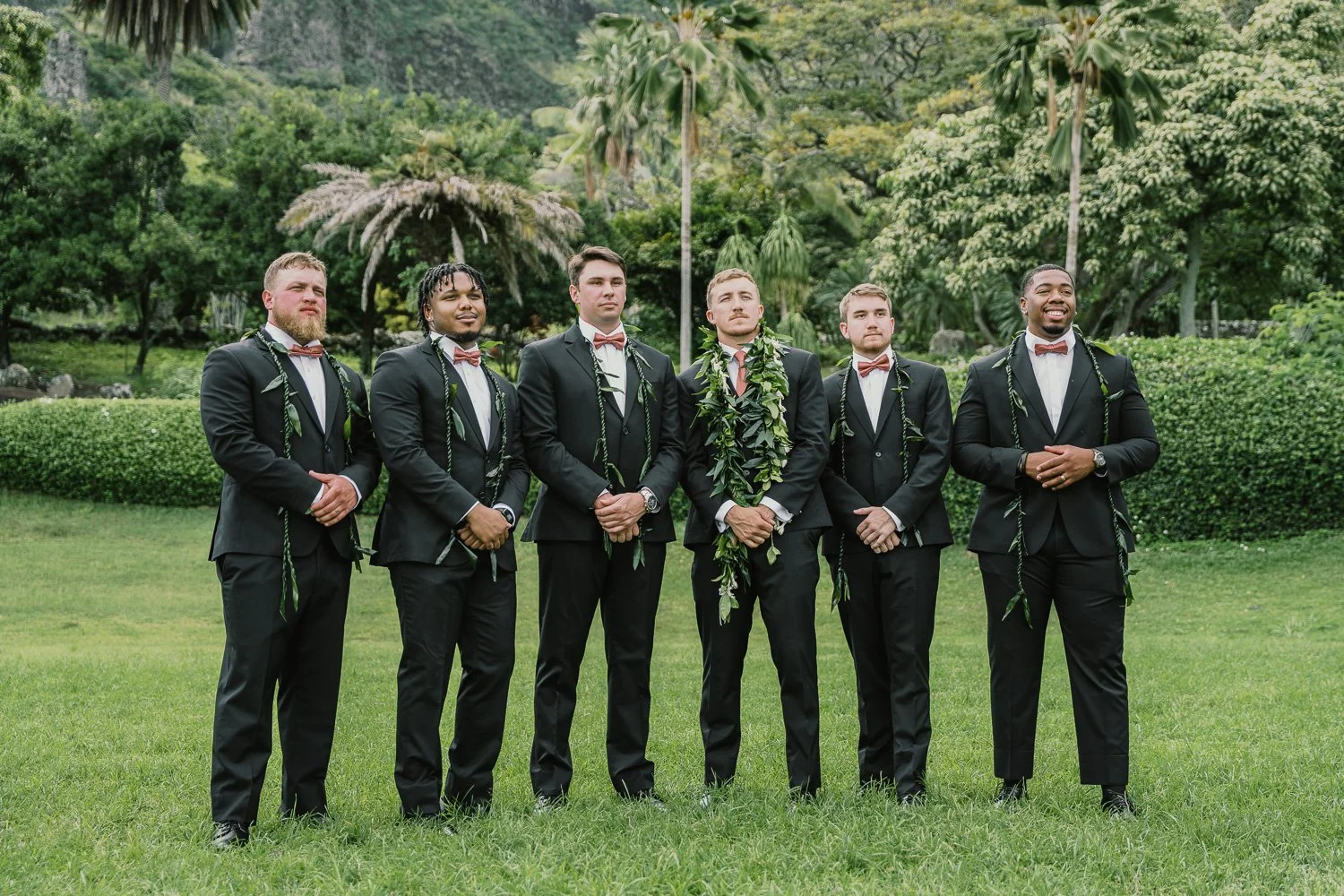 Seven men in black tuxedos and bow ties standing outdoors on a grassy lawn, with tropical trees and plants in the background, participating in a wedding ceremony.