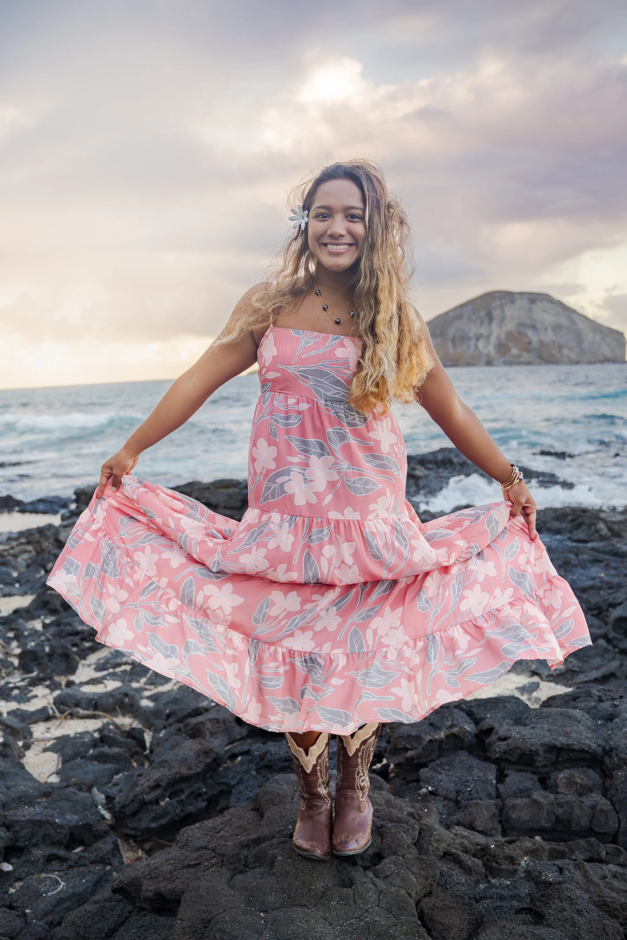 A woman in a pink floral dress standing on black rocks by the ocean, smiling, with an island in the background and a cloudy sky.