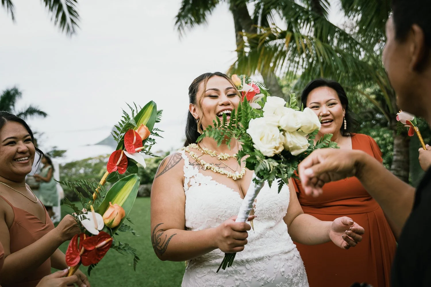 A bride in a white wedding dress holding a bouquet smiling with two women around her, exchanging floral leis during an outdoor wedding celebration under palm trees.