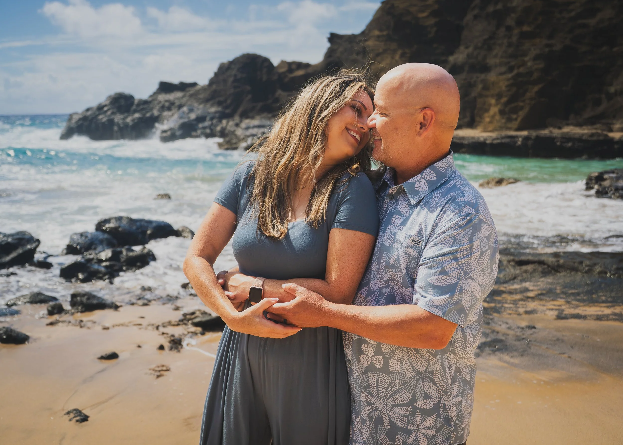 A couple enjoying a romantic moment on the beach, holding hands, with ocean waves and rocky cliffs in the background.