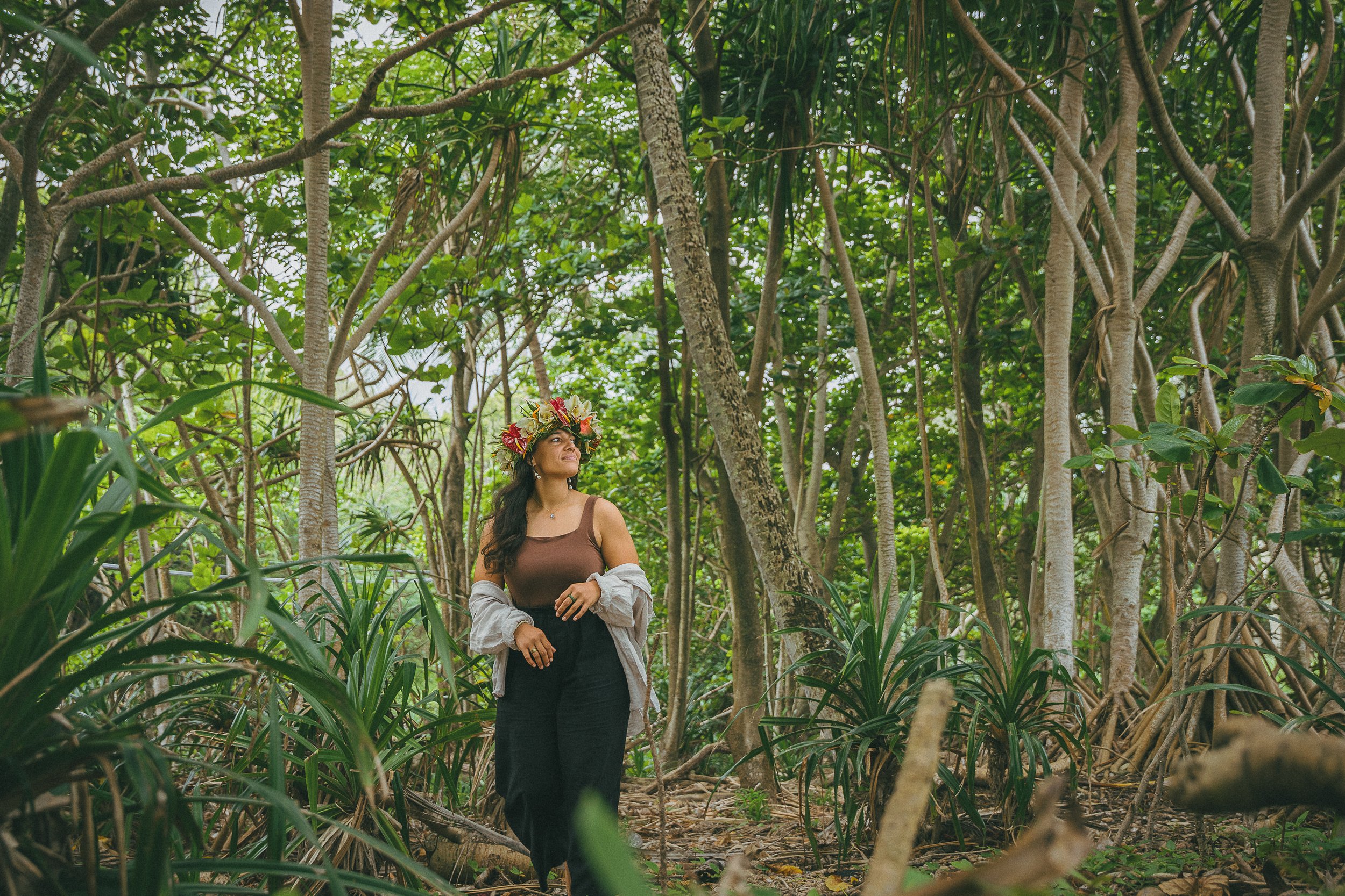 A woman wearing a flower crown stands in a dense green jungle with tall trees and lush foliage.