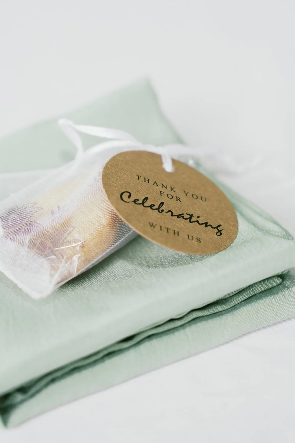 A small clear gift bag with a gold candle inside, placed on a light green cloth, with a round brown tag that says 'Thank you for celebrating with us' attached with a white ribbon.