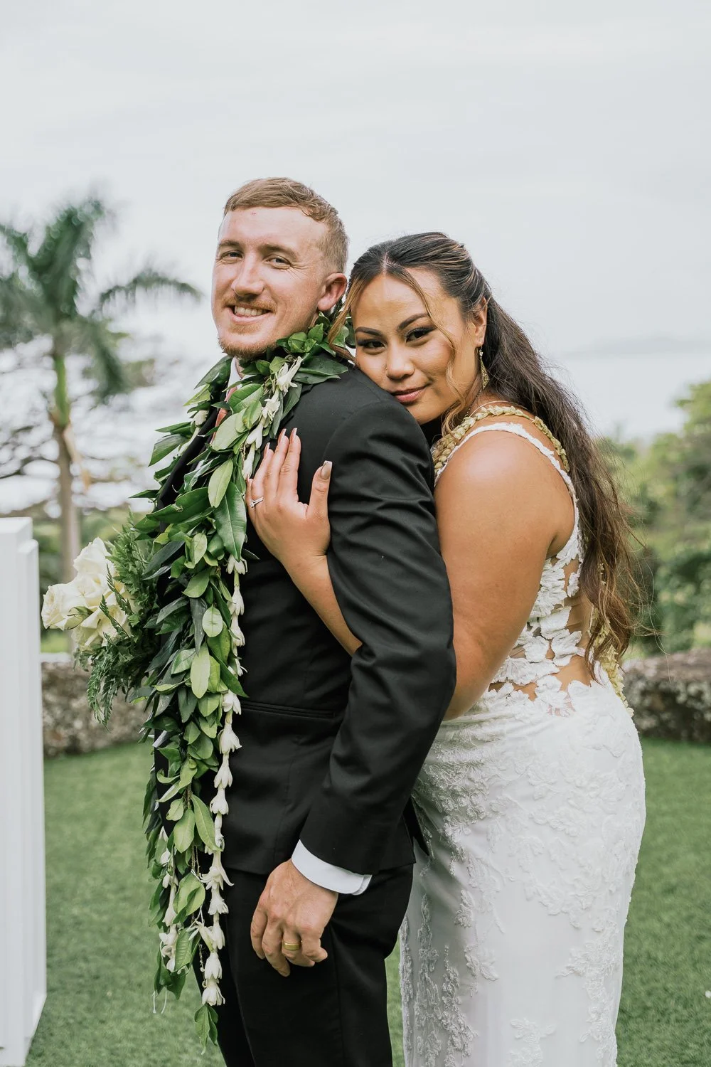 A newlywed couple, the groom wearing a black suit and lei, and the bride in a white lace wedding dress, embracing outdoors with greenery and palm trees in the background.