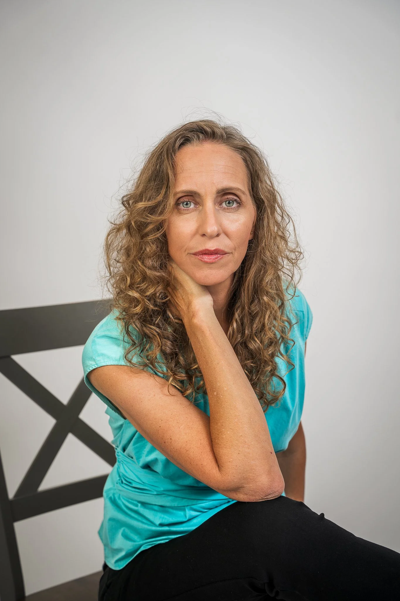 A woman with curly light brown hair, wearing a turquoise top, sitting on a black chair with a crossing pattern, resting her chin on her hand, looking at the camera with a neutral expression, against a plain white background.