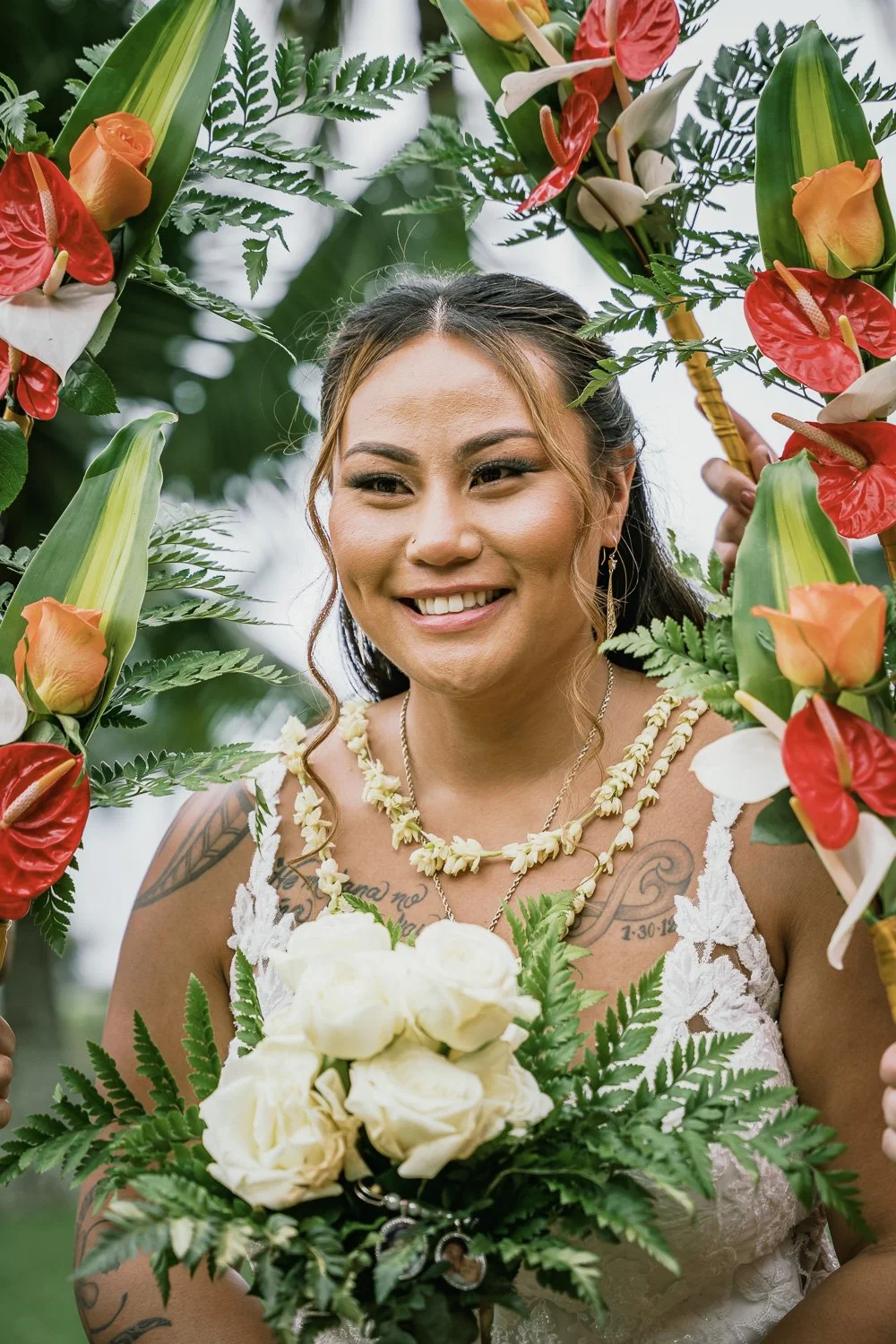 A smiling woman holding a bouquet of white roses, surrounded by a floral arch with red and orange flowers and green foliage, outdoors in a tropical setting.