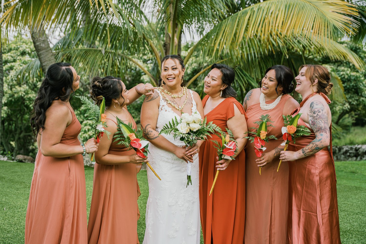 A bride in a white wedding dress holding a bouquet, surrounded by five bridesmaids in burnt orange dresses, all smiling and holding flowers, outdoors with lush green trees in the background.
