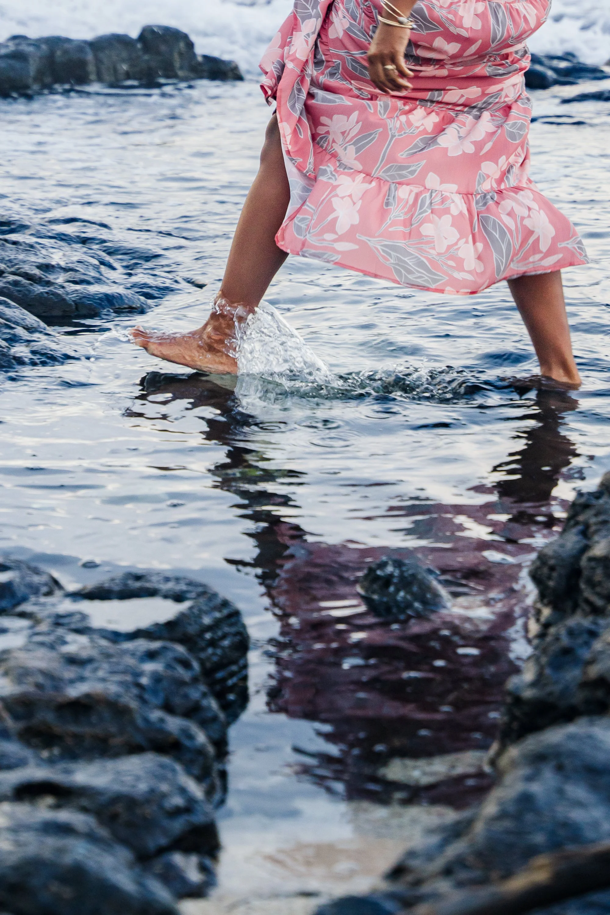 Person walking in shallow ocean water, wearing a pink floral dress, with rocks visible in the foreground.