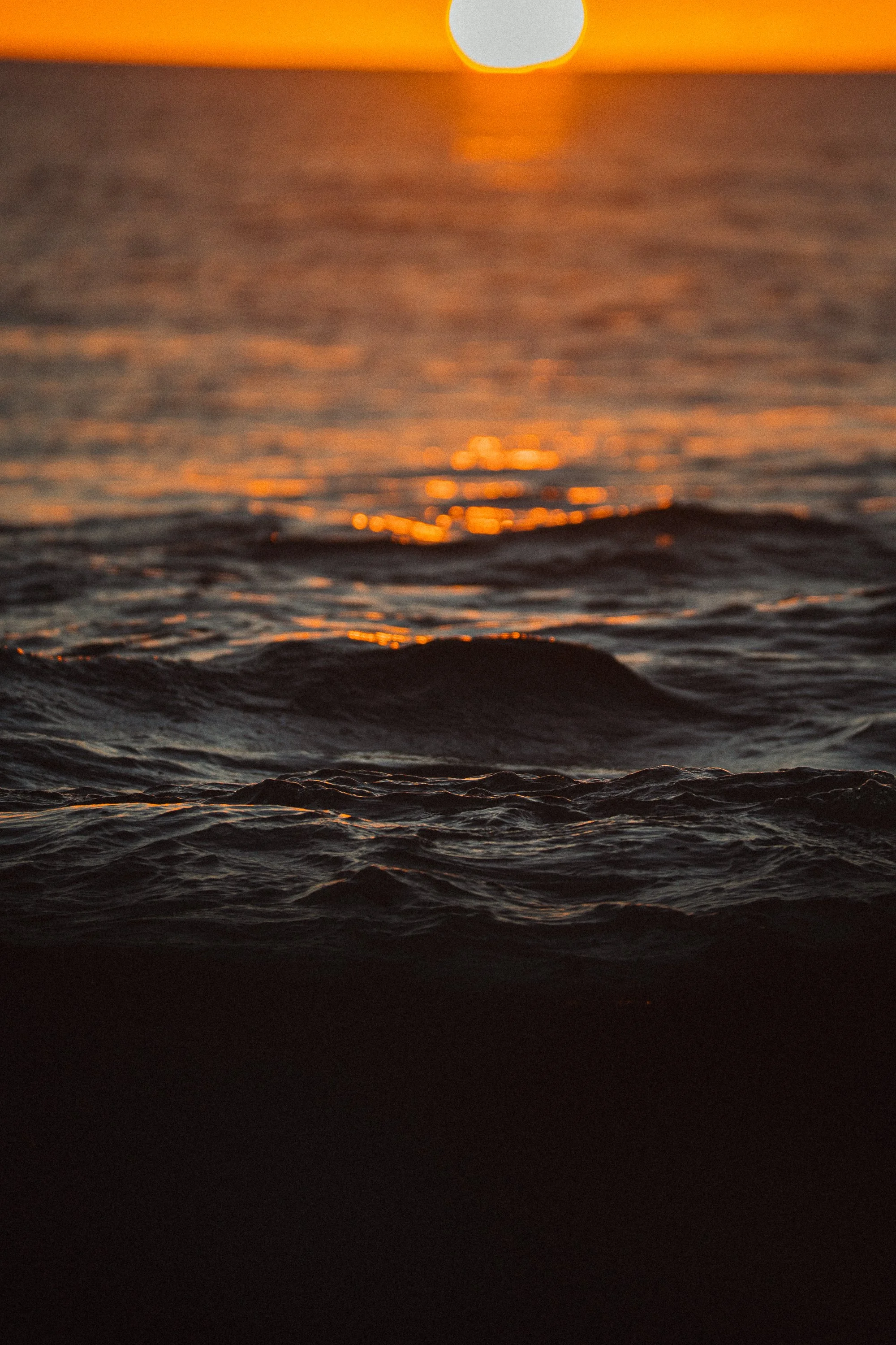 A close-up view of ocean waves during sunset, with the sun low on the horizon and the sky glowing orange.