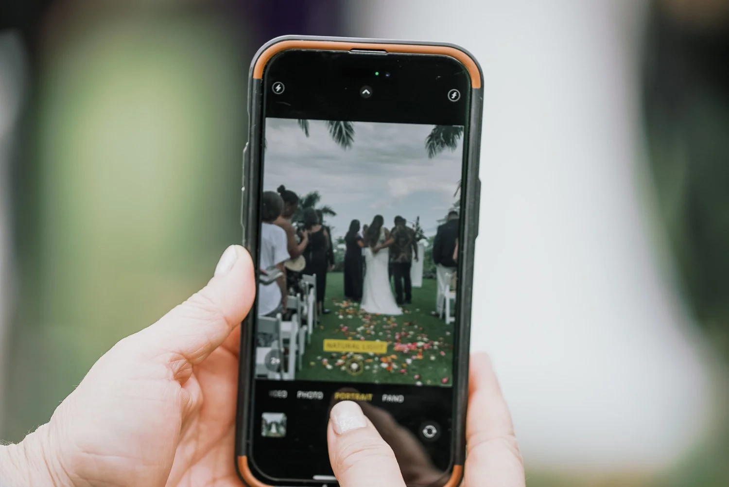 Person taking a photo of a wedding ceremony on a smartphone. The wedding party is visible through the phone's screen, with a couple dancing or hugging outdoors on a grassy area decorated with flower petals and chairs, under a cloudy sky with palm tre