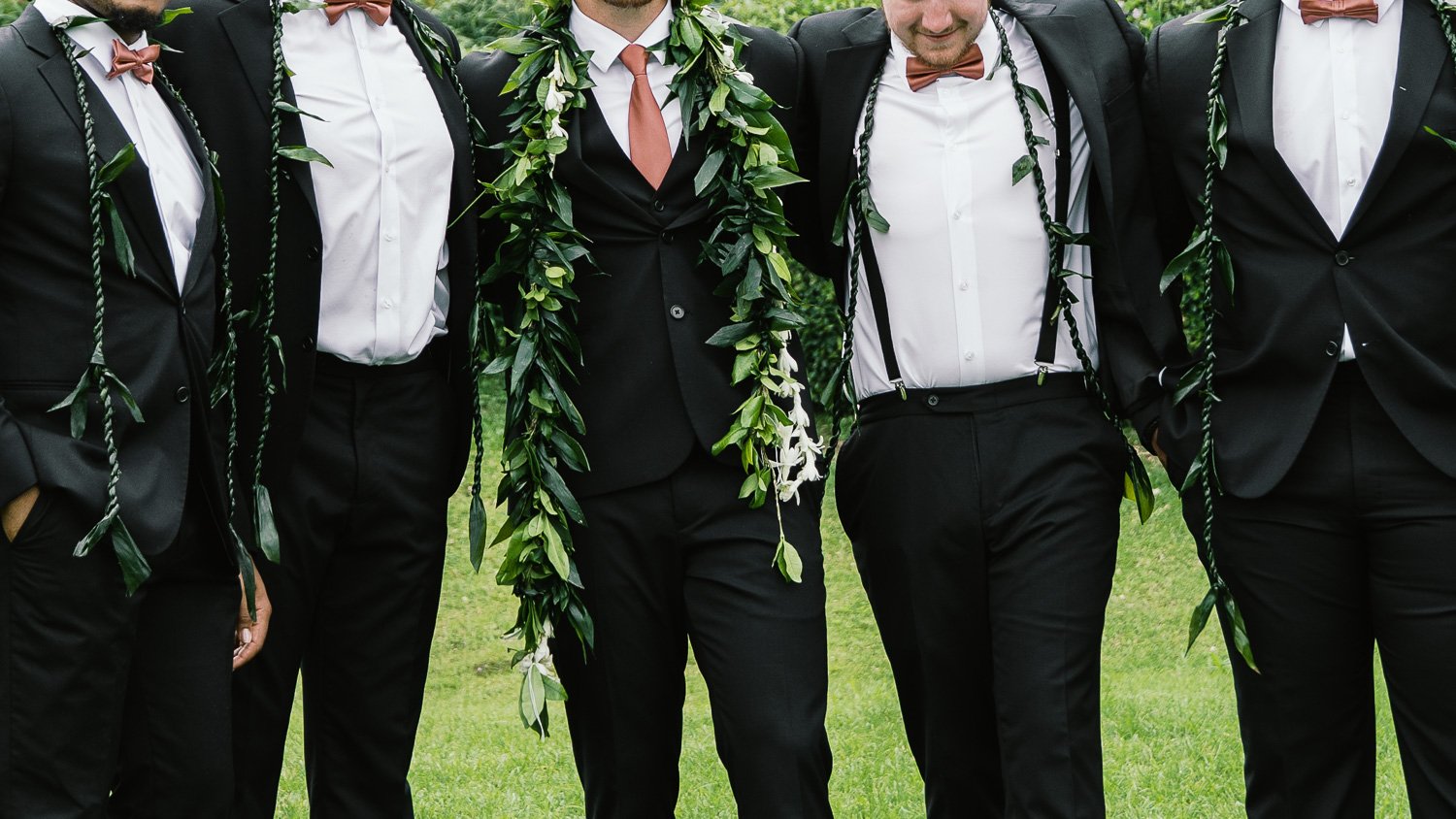 Close-up of a group of men dressed in black suits with white shirts and brown bow ties, standing outdoors on grass, with one person wearing a lei made of green leaves around their neck.