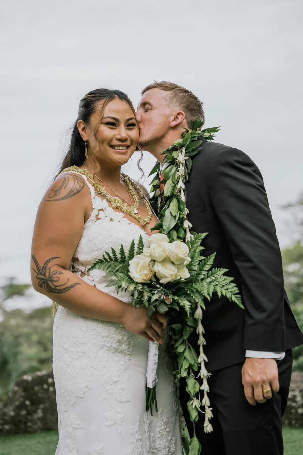 A newlywed couple on their wedding day, the groom is kissing the bride's cheek while she smiles, holding a bouquet of white roses and greenery, outdoors against a cloudy sky.
