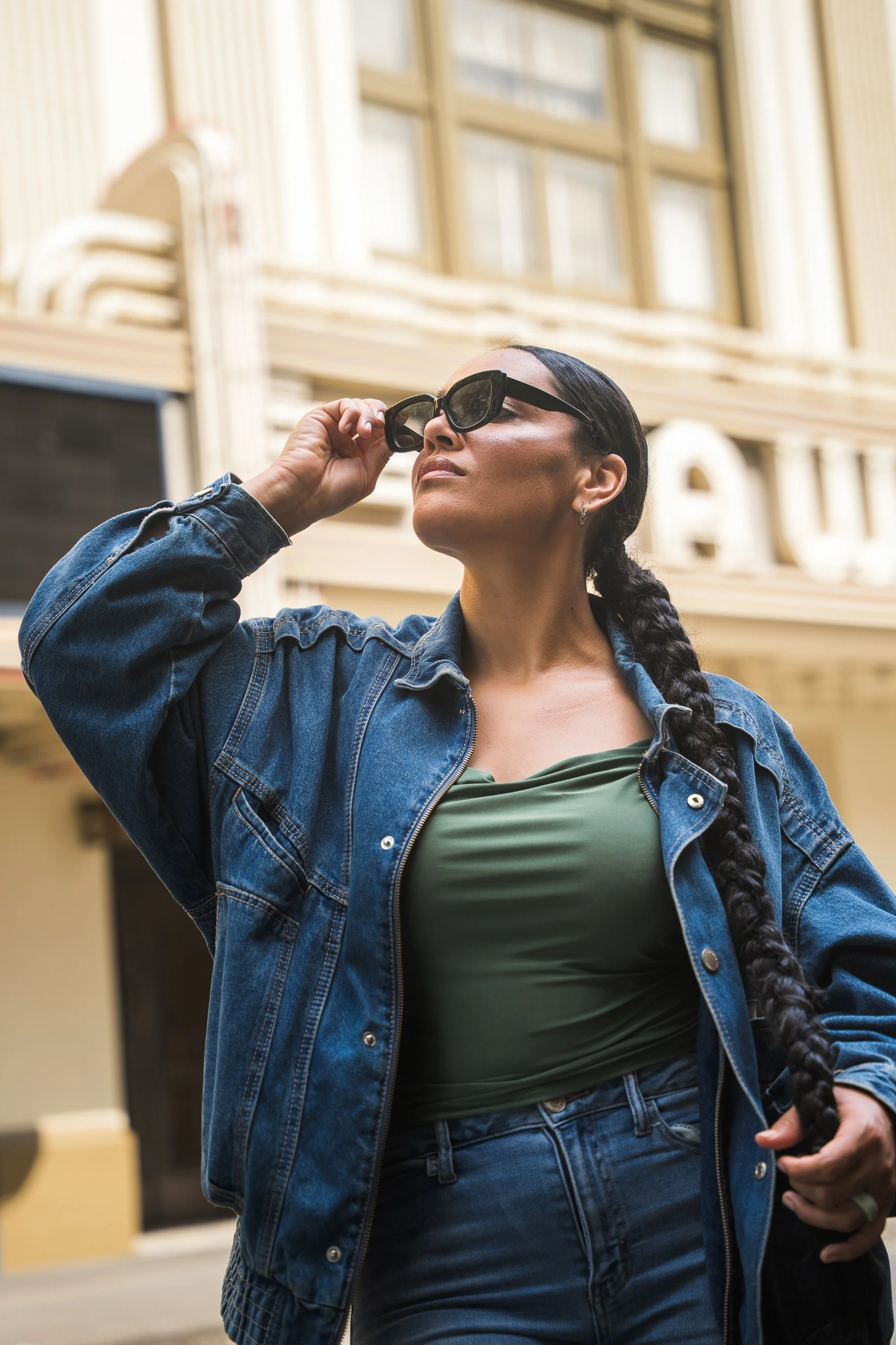 A confident woman with a braid and sunglasses adjusting her glasses outdoors, wearing a denim jacket and green top.