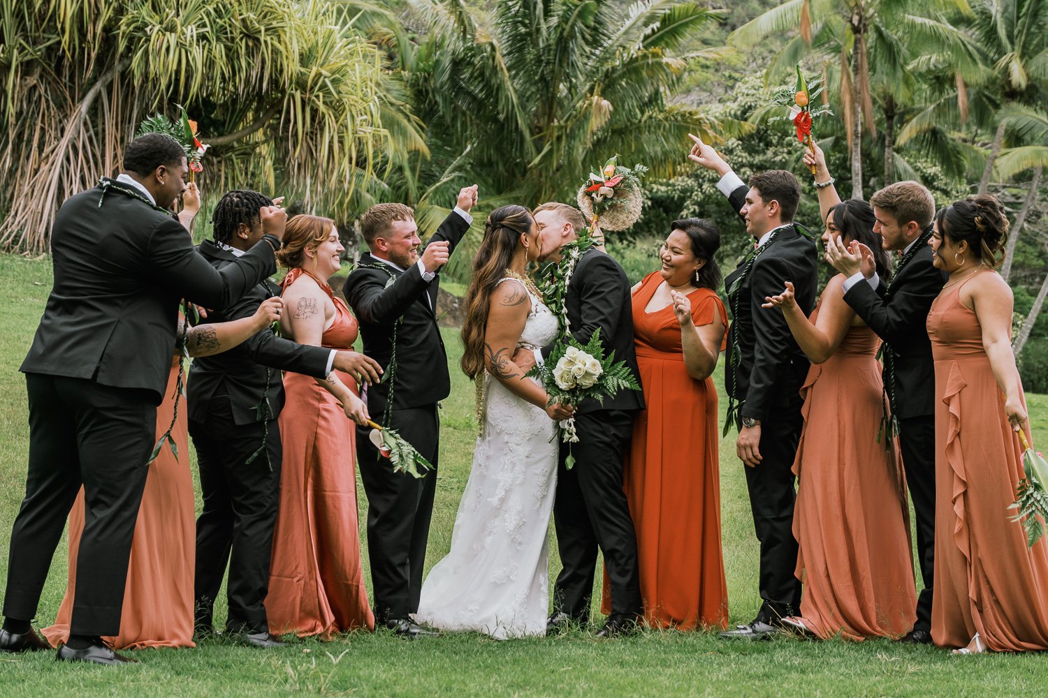 A wedding celebration outdoors with the couple kissing, surrounded by wedding party members cheering and clapping in a lush green garden.