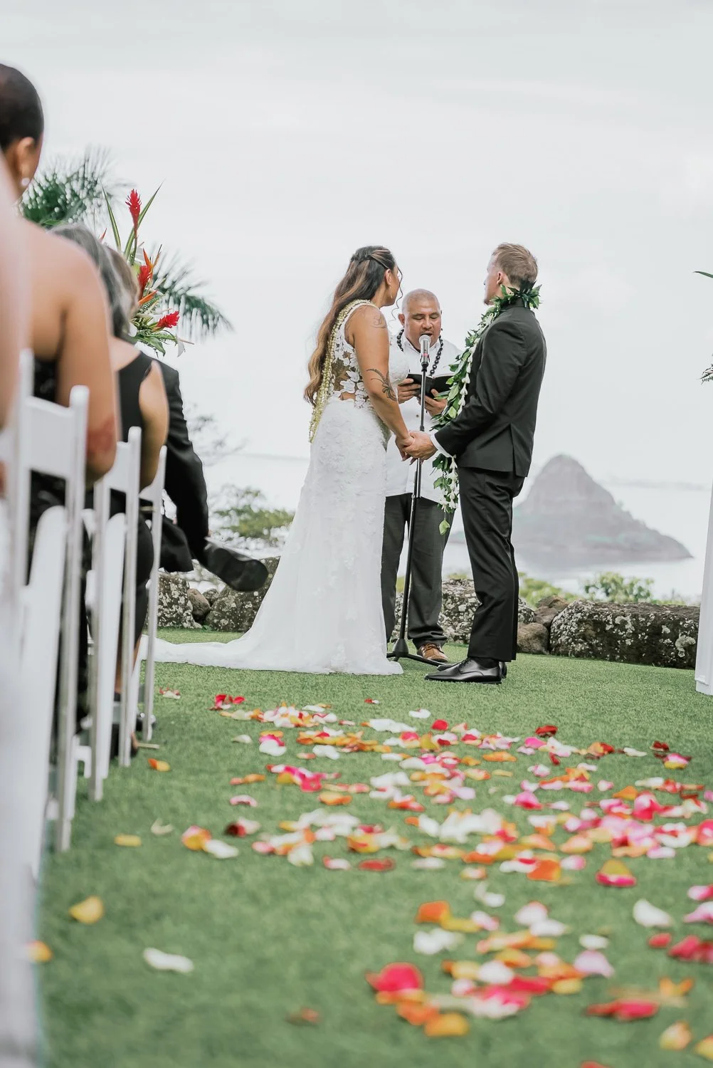A couple getting married outdoors, holding hands and exchanging vows, with a ceremonial officiant reading in front of them, on a grassy area decorated with rose petals and tropical scenery in the background.