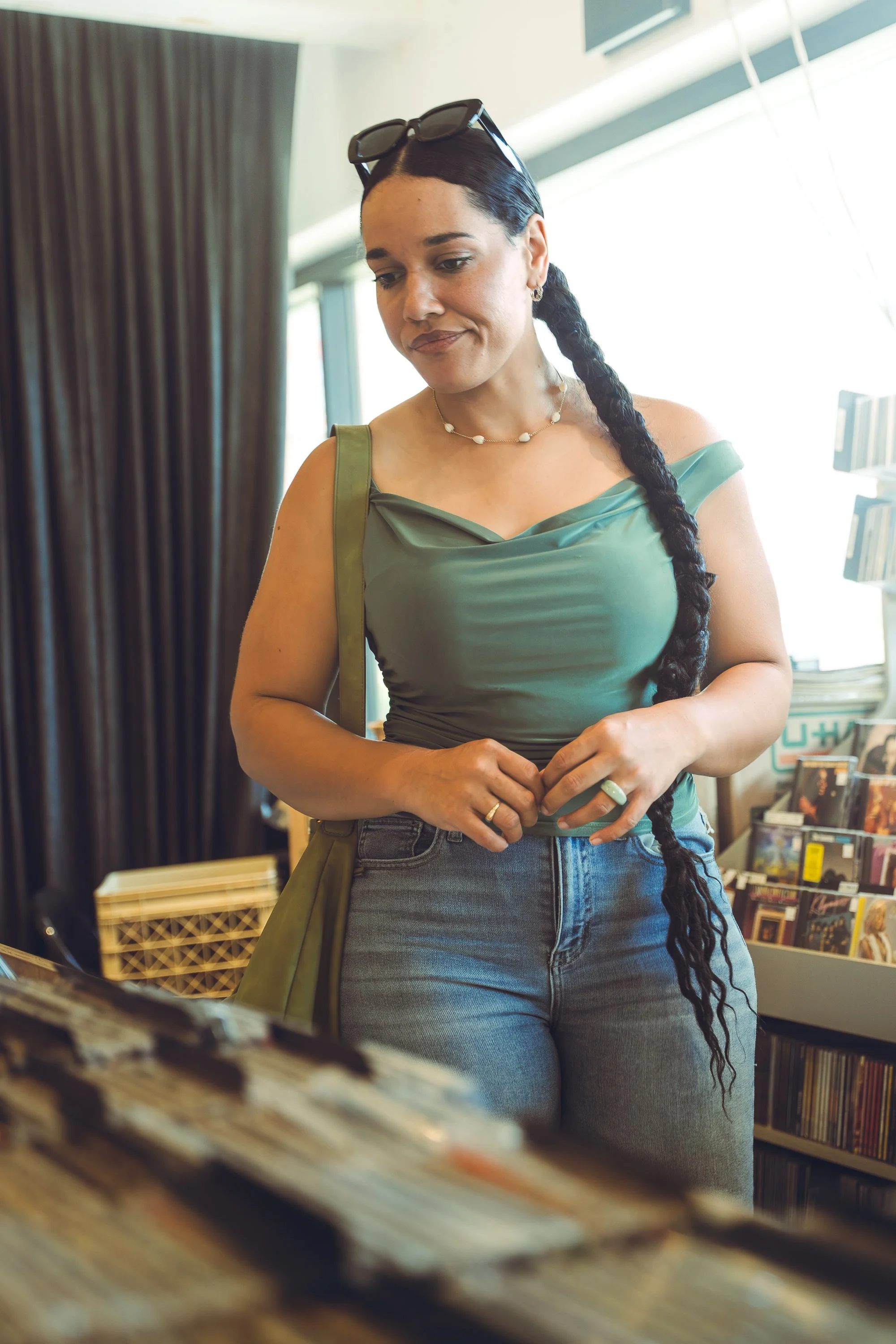 A woman with long dark hair in a braid, wearing a green top, jeans, and sunglasses on her head, shopping in a record store.