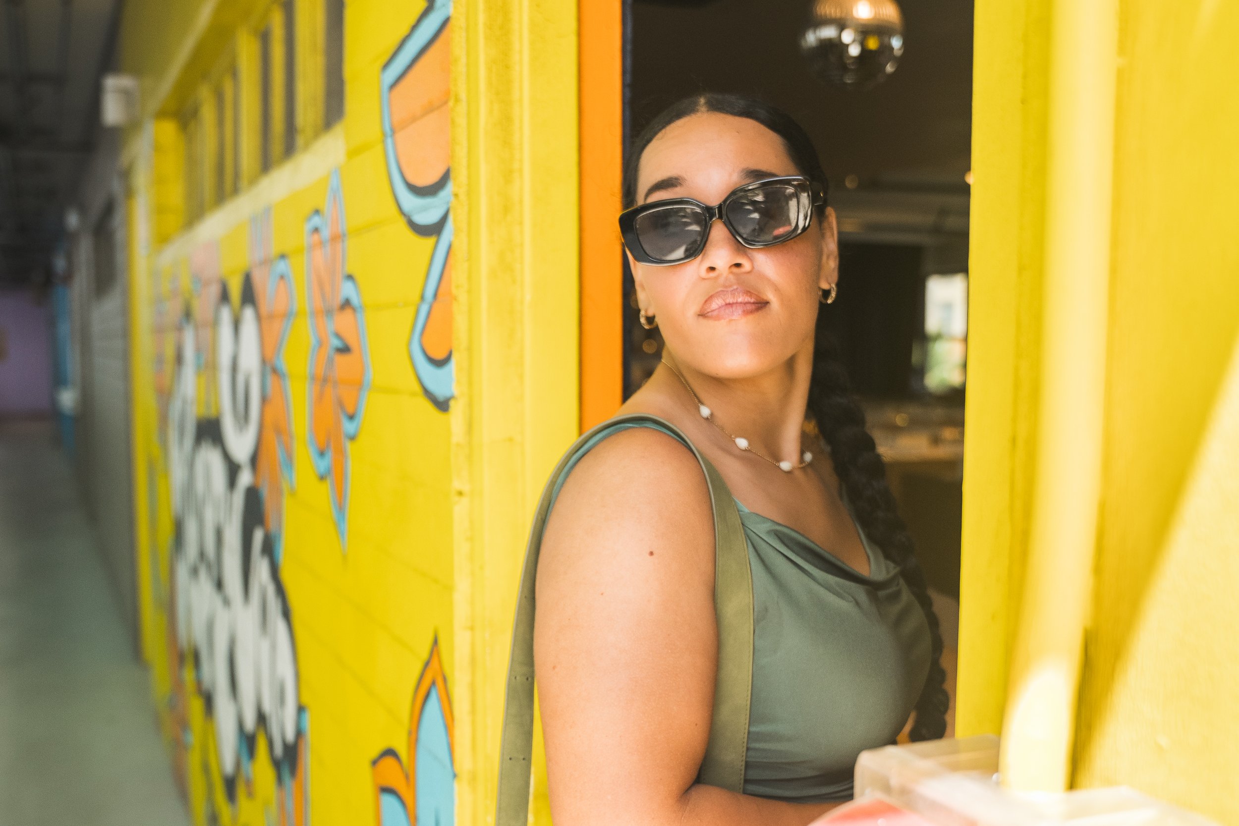 A woman wearing sunglasses and a green dress standing near a vibrant yellow wall with colorful abstract graffiti art.
