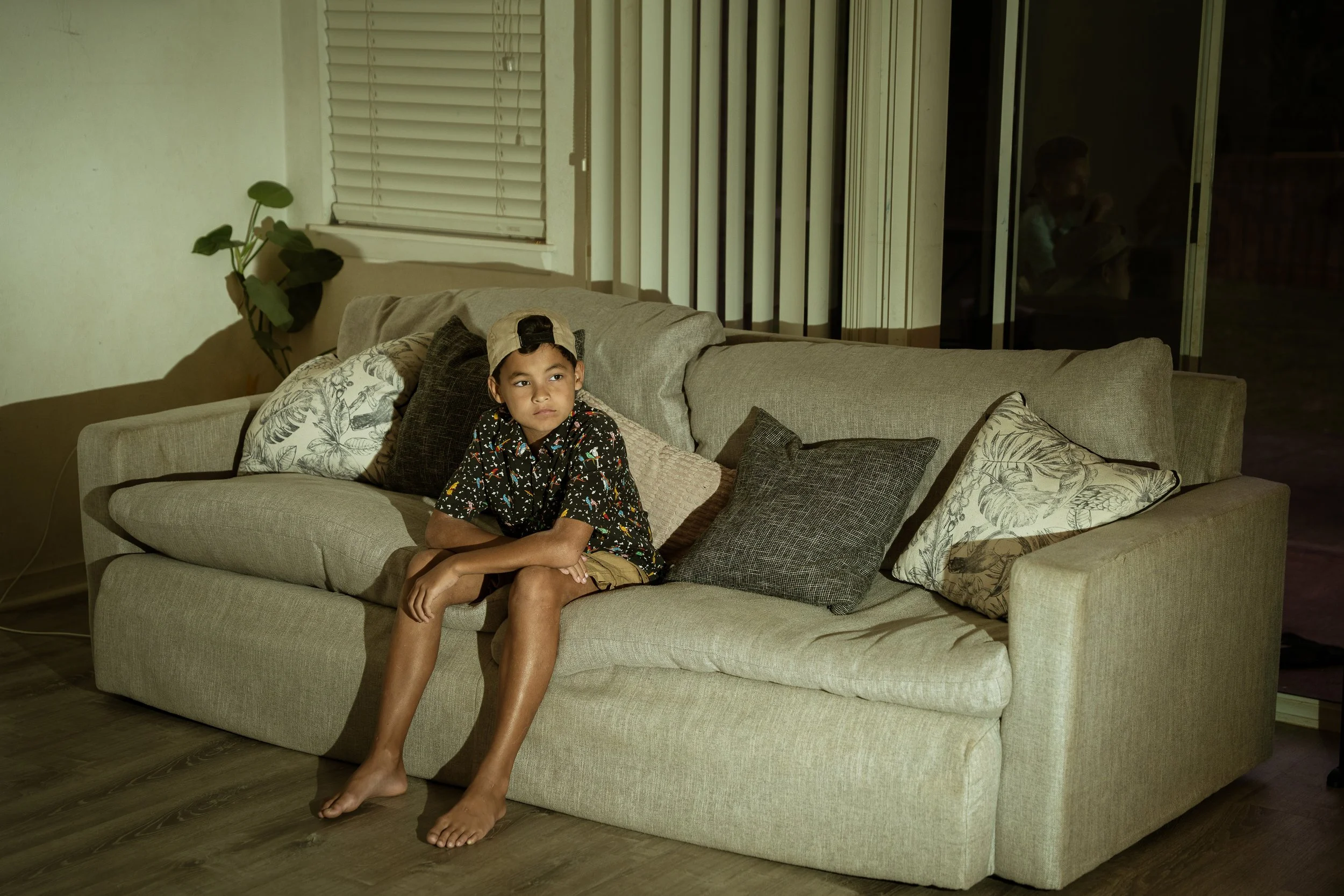 Boy sits and watches tv. His brother reflection is caught in the reflection of the window.