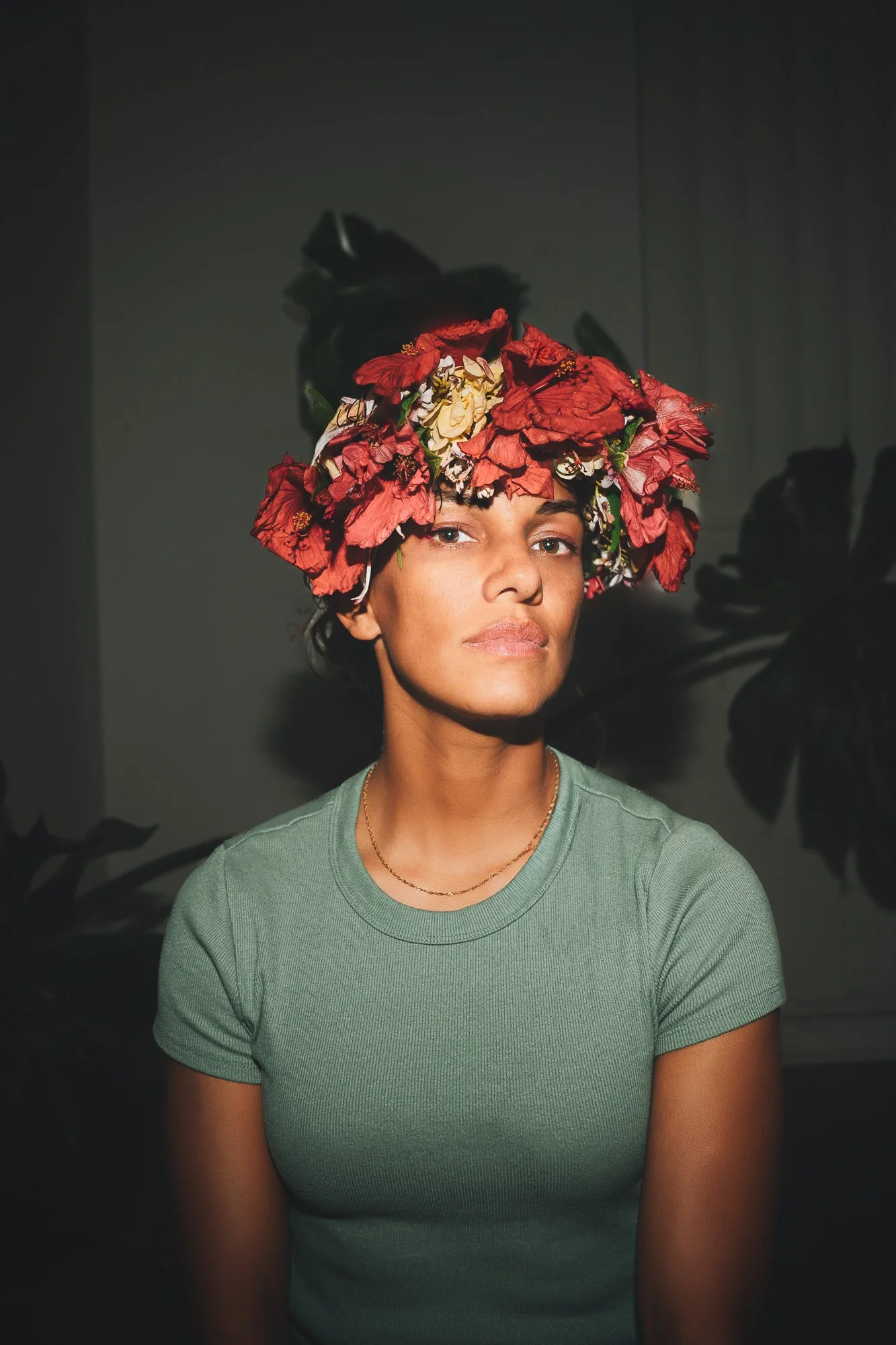 A woman with short dark hair wearing a floral headdress of red and cream flowers, a green t-shirt, and a delicate gold necklace. She is looking at the camera with a neutral expression, and dark foliage is visible in the background.