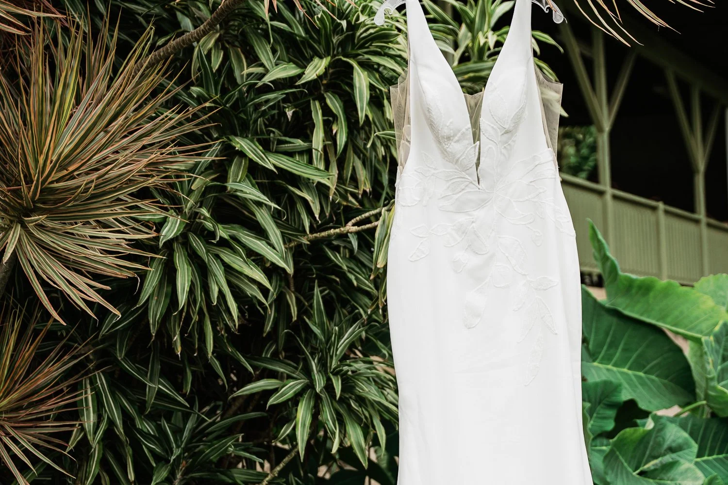 A white wedding dress with floral embroidery hanging outdoors amidst lush green tropical plants.