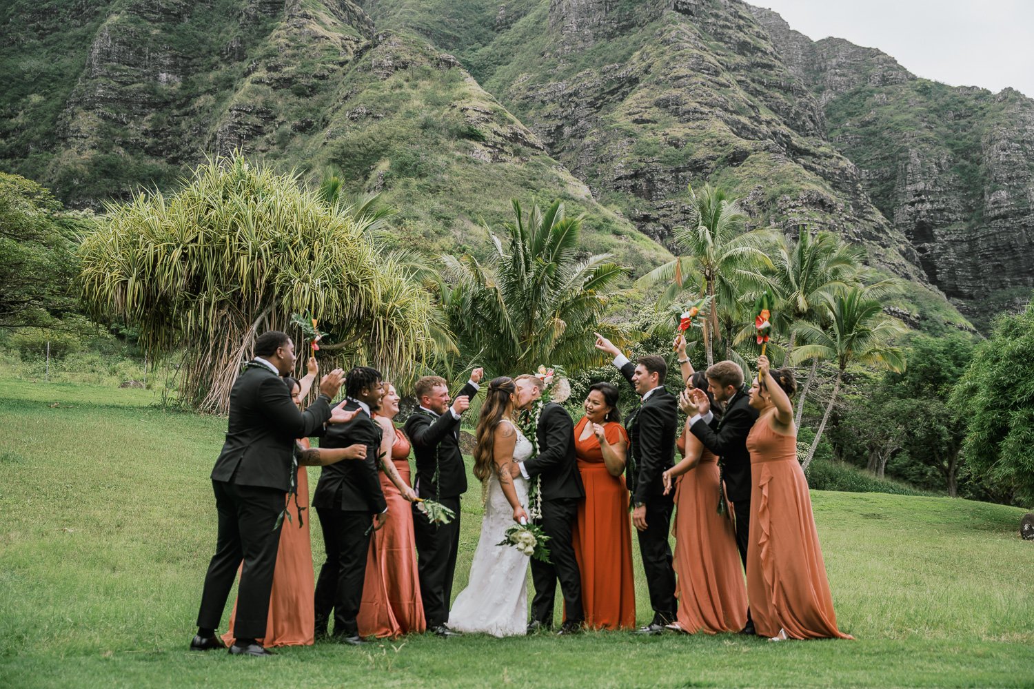 A wedding party of four men and six women, with the bride and groom in the center, celebrating outdoors in a lush green landscape with mountains in the background. The women are wearing orange gowns or peach dresses, and the men are in black suits. T