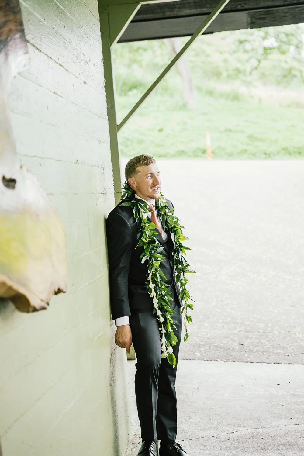 Young man in a suit with a leafy lei, standing against a green wall under a shelter, smiling and looking to the side.