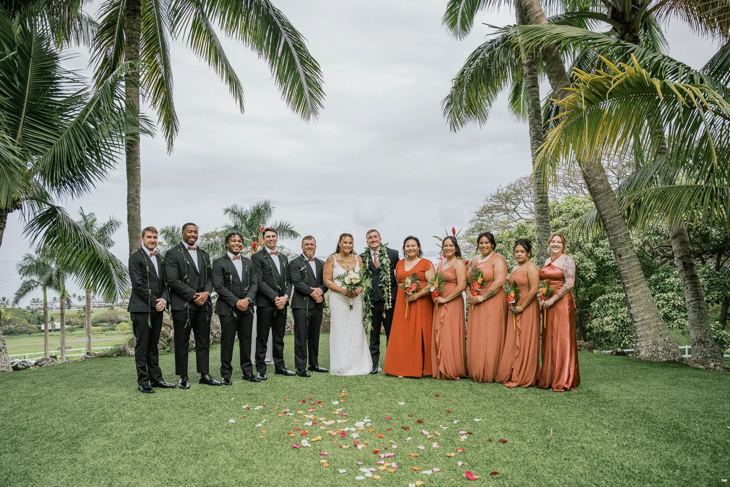 Wedding party standing outdoors on grass with palm trees and cloudy sky in background, with scattered flower petals on the ground.