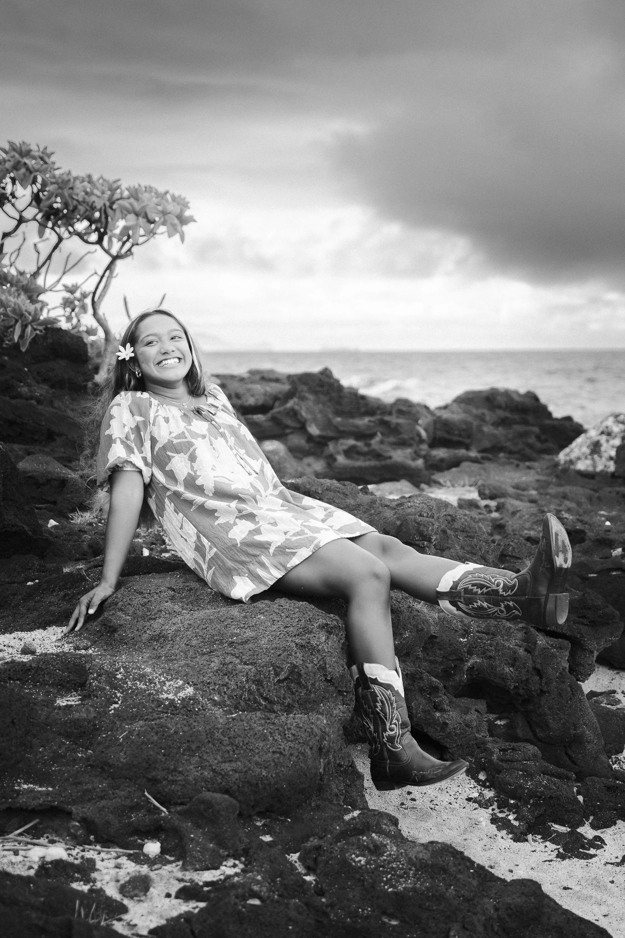 A young girl wearing cowboy boots and a floral dress is sitting on rocks by the beach, smiling with a flower in her hair, with the ocean and cloudy sky in the background.
