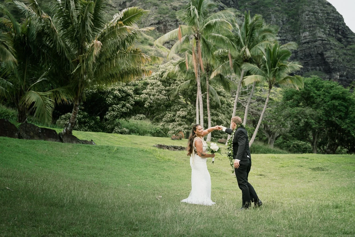 A bride and groom holding hands in a lush tropical garden with palm trees and green hills in the background, dressed in wedding attire.