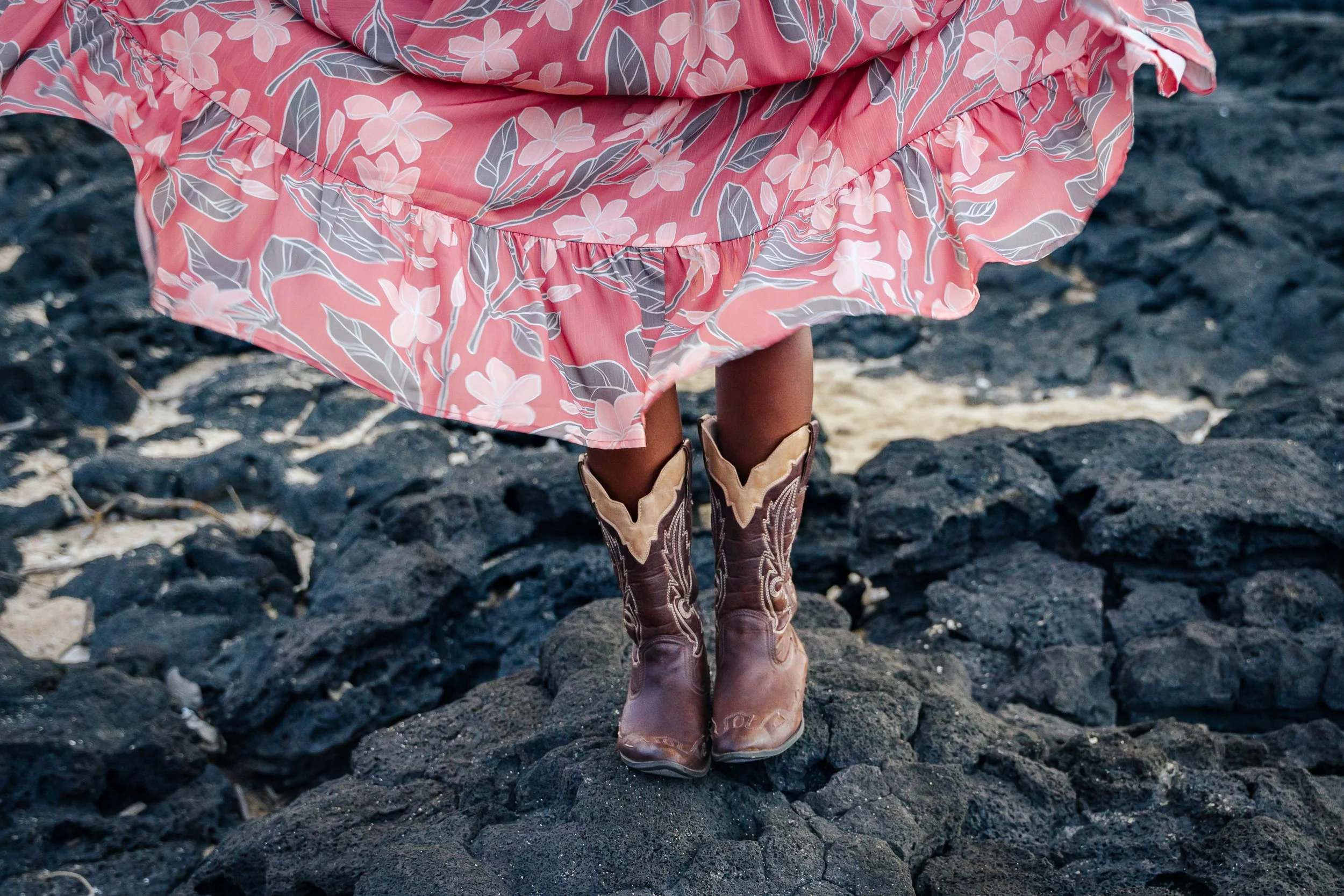 Person standing on black volcanic rocks, wearing a pink floral dress and cowboy boots.